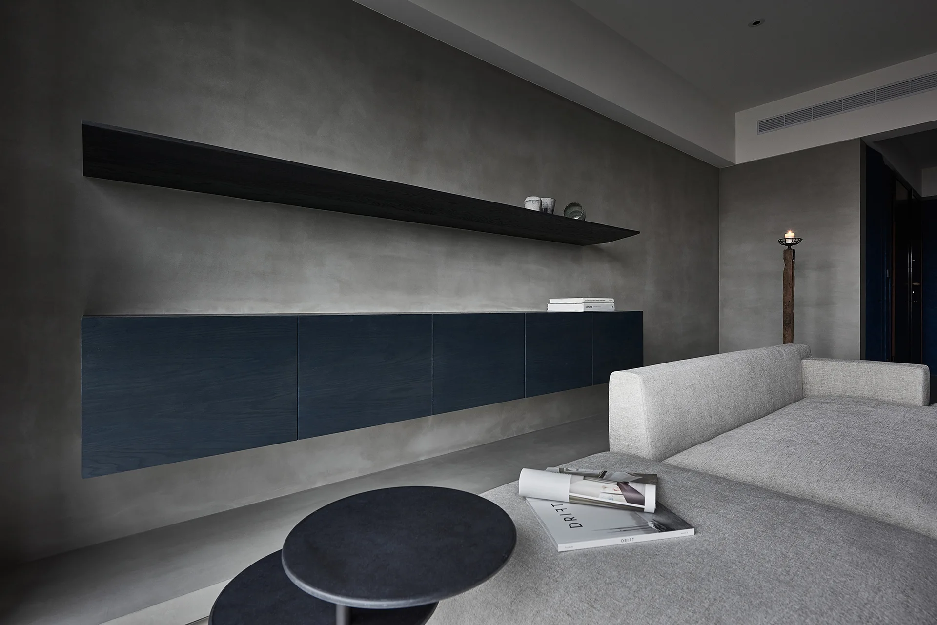 A minimalist living room features a grey upholstered sofa next to a concrete-textured wall with a floating dark blue cabinet and long black shelf.