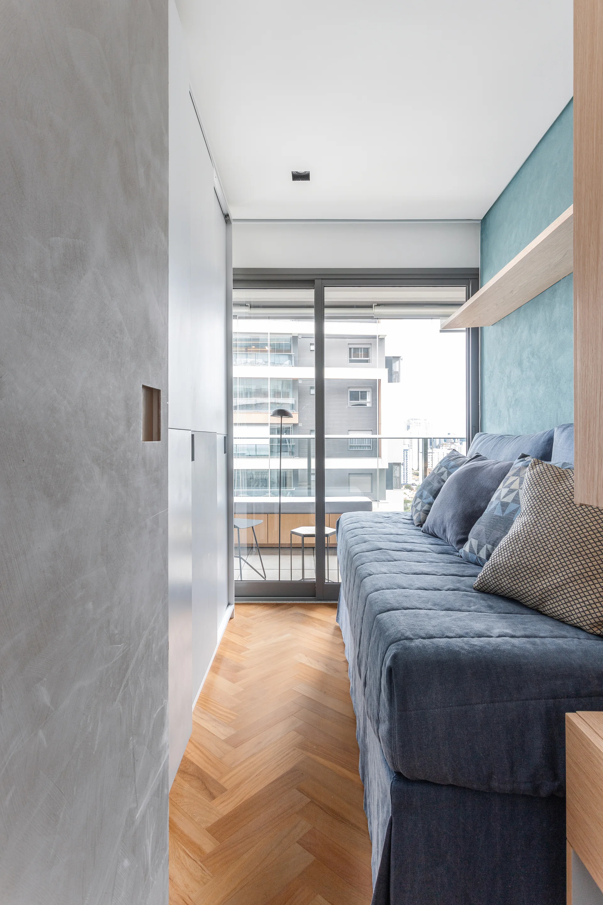 A narrow bedroom features a blue daybed against a blue accent wall, grey built-in closets, and herringbone wood flooring leading to a balcony door.