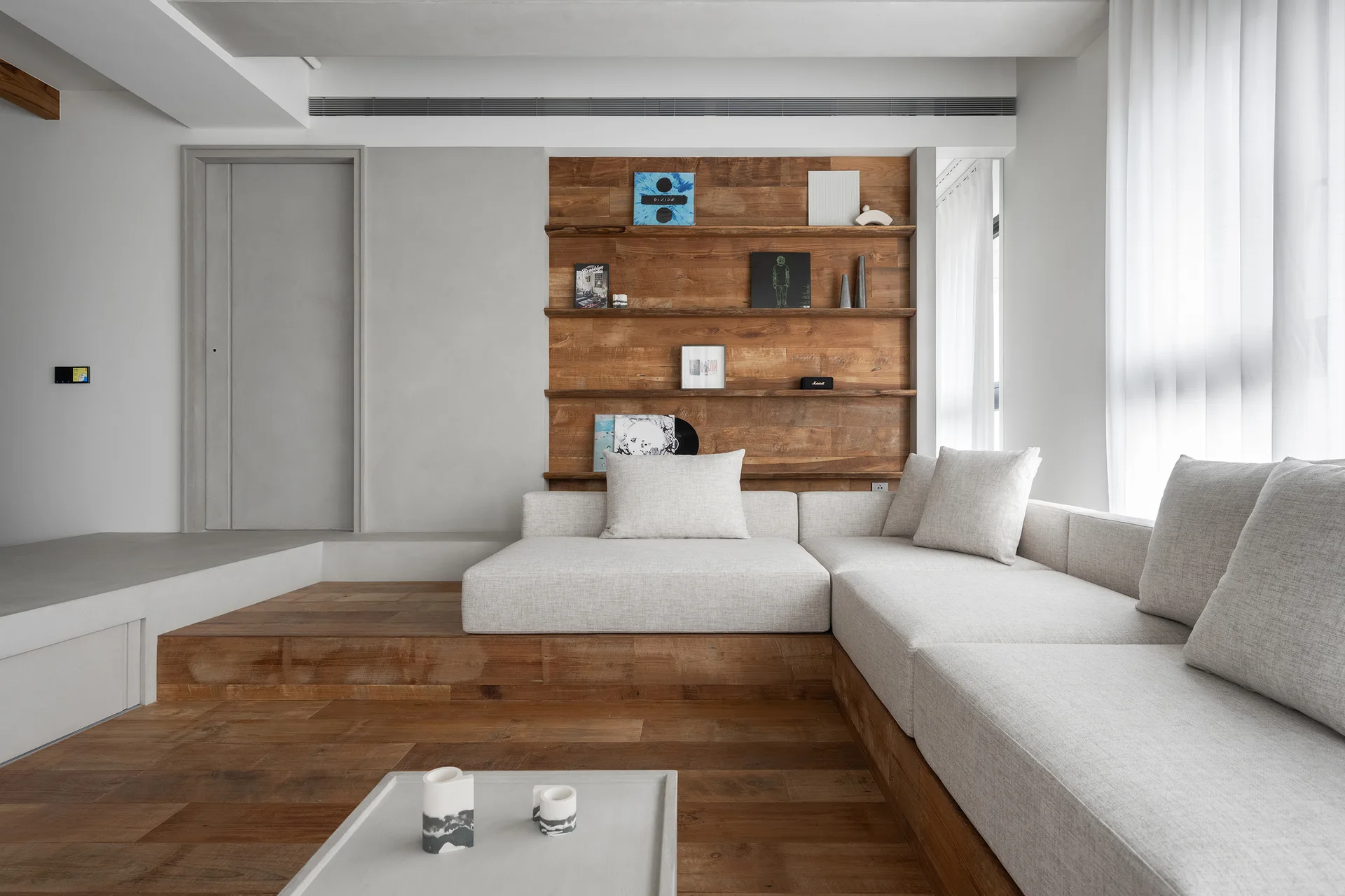An off-white sectional sofa sits on a raised wood platform floor next to a matching wood wall niche with open shelving in a minimalist room.