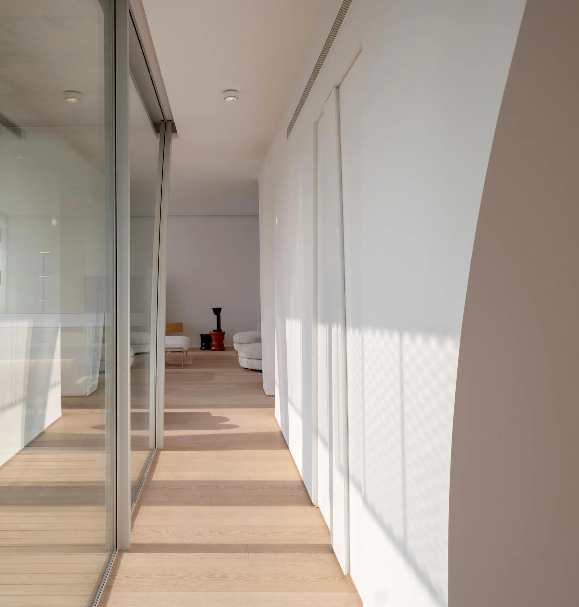 A minimalist hallway features a floor-to-ceiling glass partition wall on one side, flush white doors on the other, and light wood plank flooring.