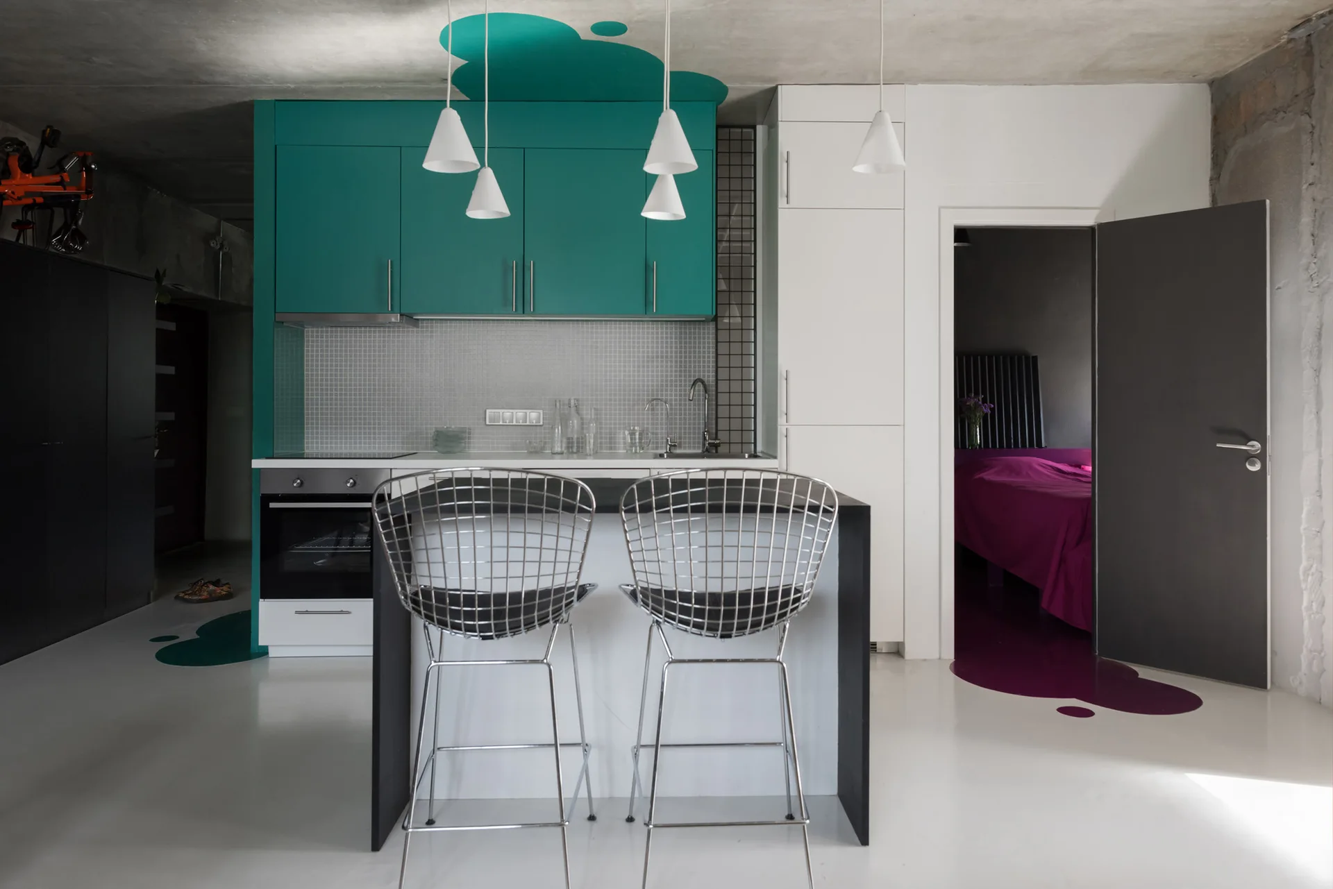 A contemporary studio kitchen featuring teal cabinets, a dark island with wire mesh bar stools, and a raw concrete ceiling above a white floor.