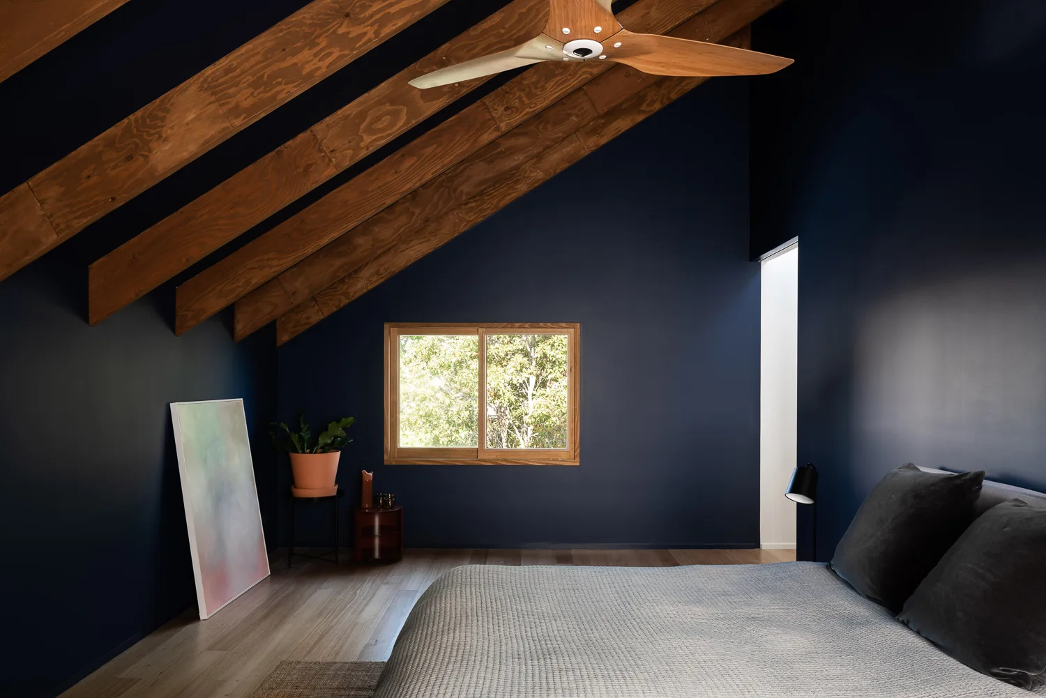 A minimalist bedroom features dark navy walls, sloped wooden ceiling beams, a matching wooden fan, and a gray bed.