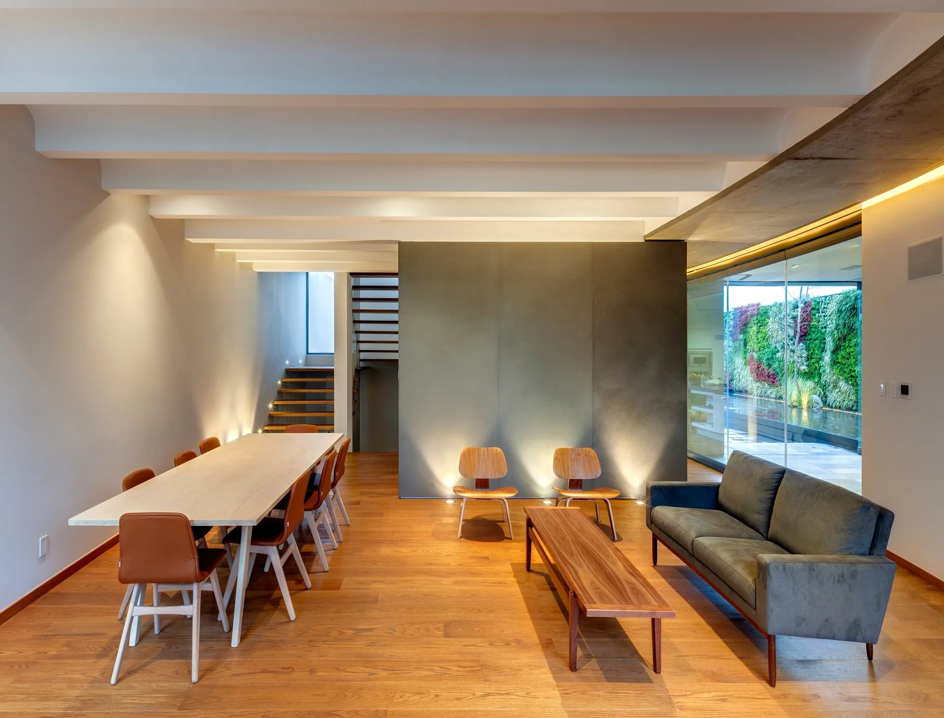 A contemporary open-plan living and dining room featuring white ceiling beams, a long wooden dining table, and a dark grey sofa.