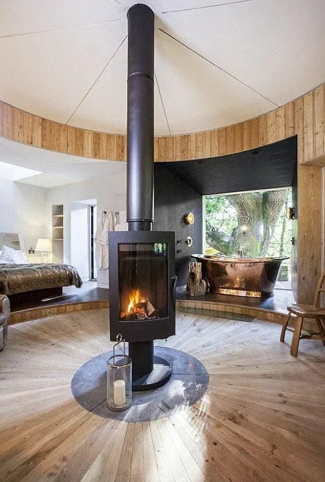 A circular bedroom and bath suite featuring a central black freestanding fireplace, radial wood flooring, and a copper bathtub by a large window.