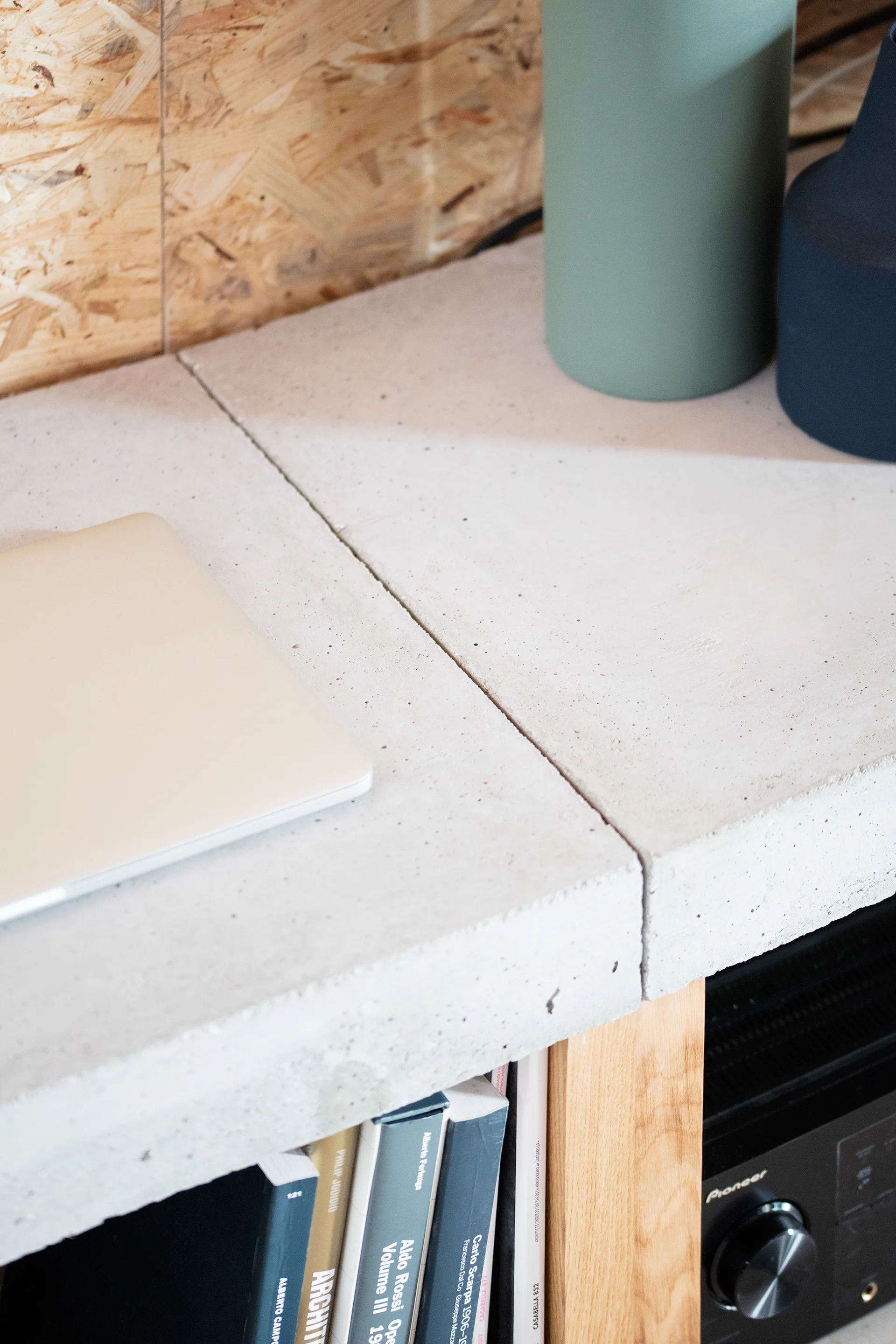 A close-up of a textured light grey concrete shelf supporting a laptop and vases, with books and an audio receiver in the wooden compartment below.