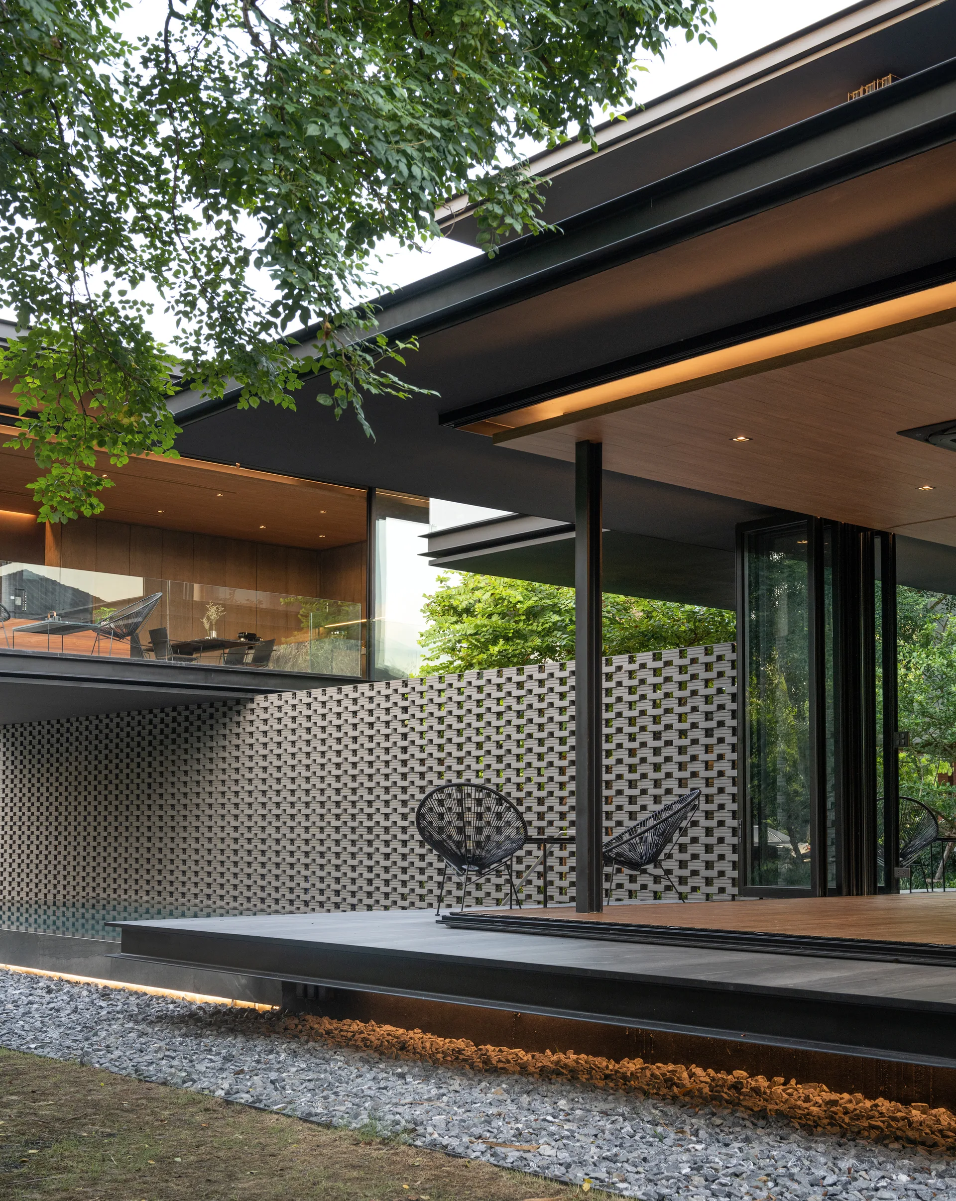 A modern covered patio features a perforated grey brick screen wall, dark wood decking, and folding glass doors next to black accent chairs.