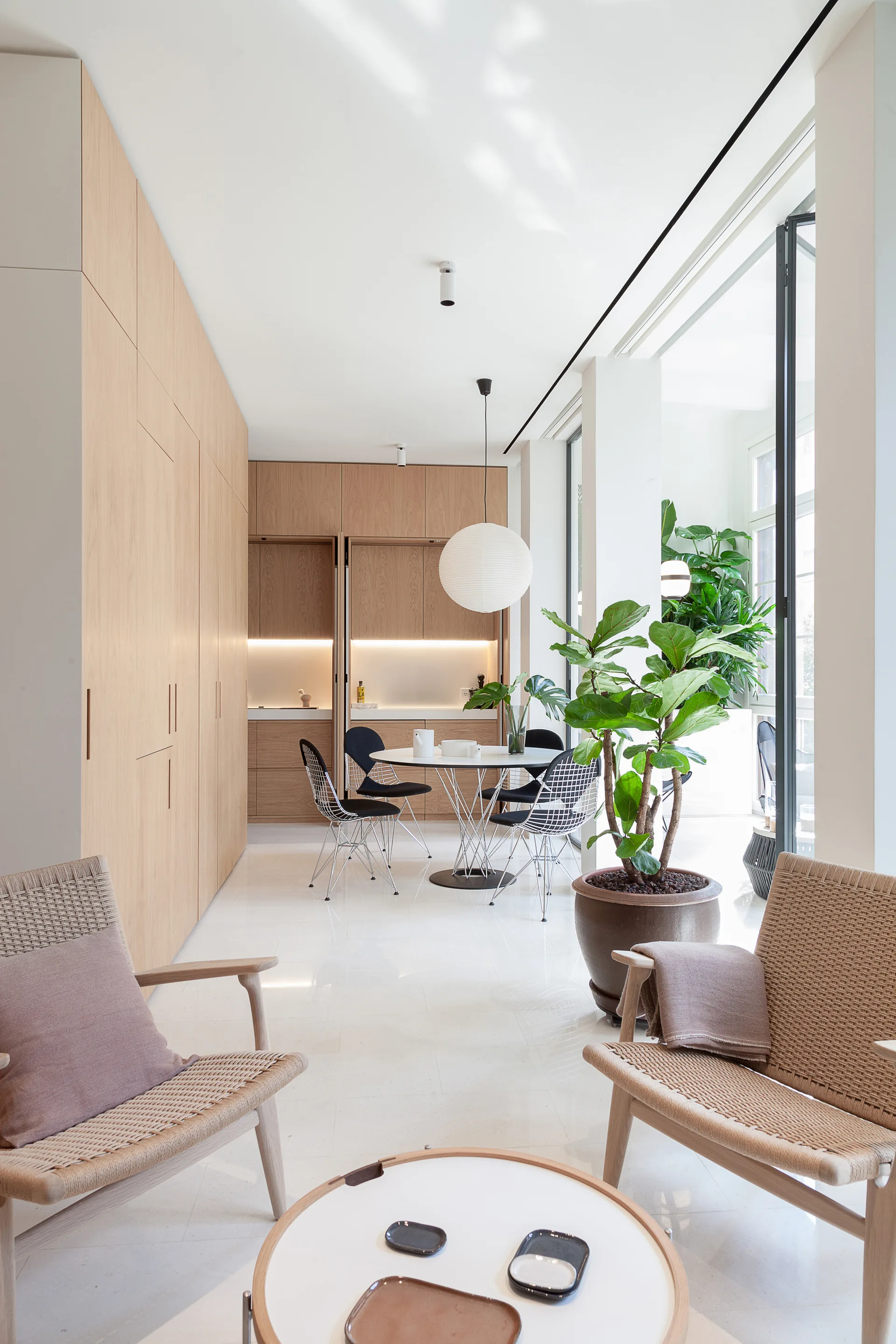 A bright open-plan room features a round dining table with wire chairs, a wall of seamless light oak cabinetry, and woven lounge chairs in the foreground.