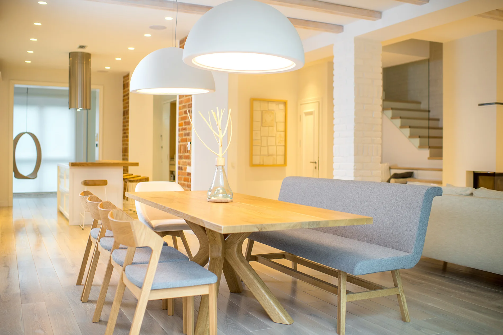 A light wood dining table features individual chairs on one side and a gray upholstered high-backed bench on the other under two white dome pendant lights.