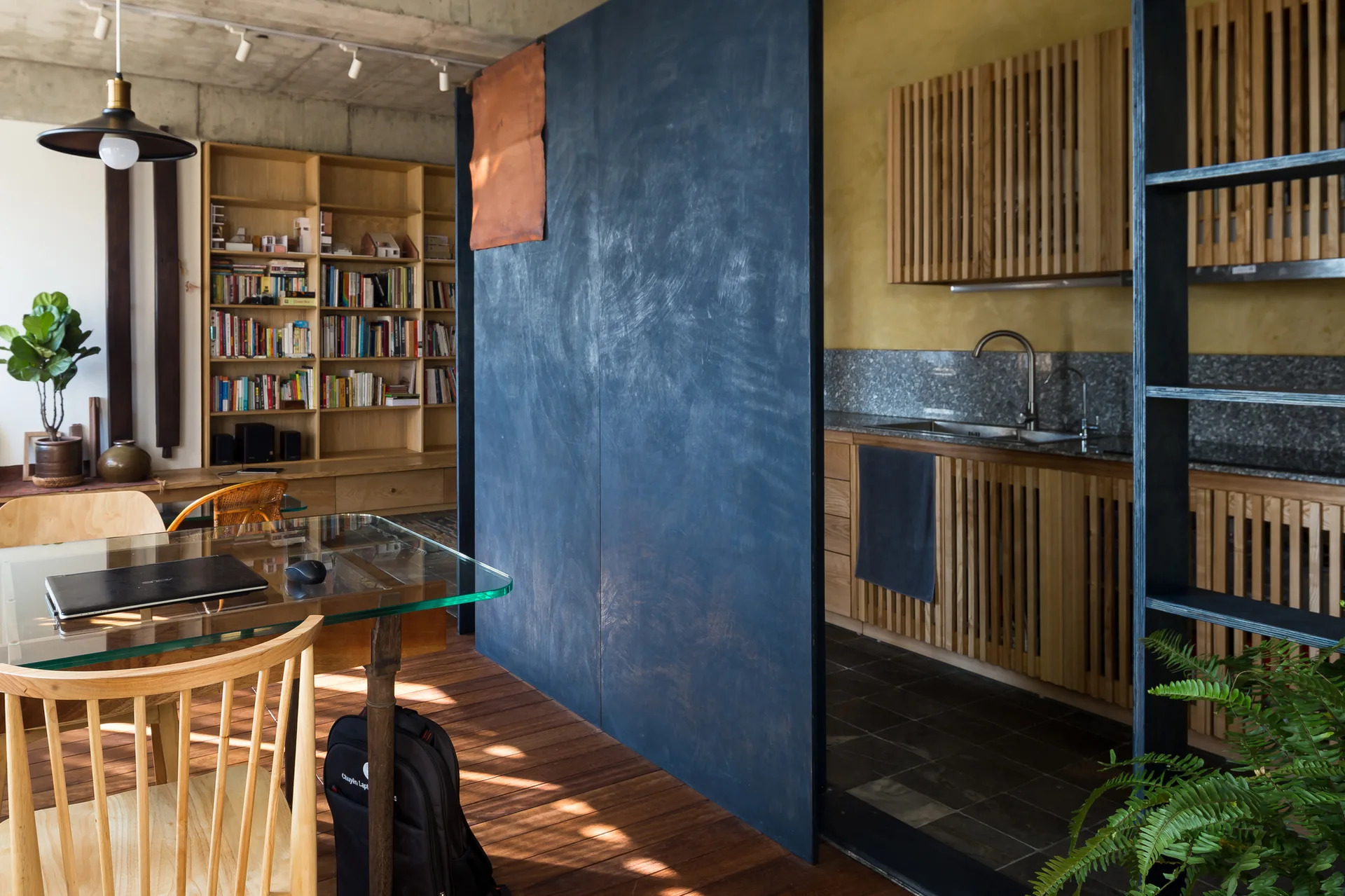 A dark blue room divider separates a kitchen with slatted wood cabinets from a dining area with a glass table and concrete ceiling.