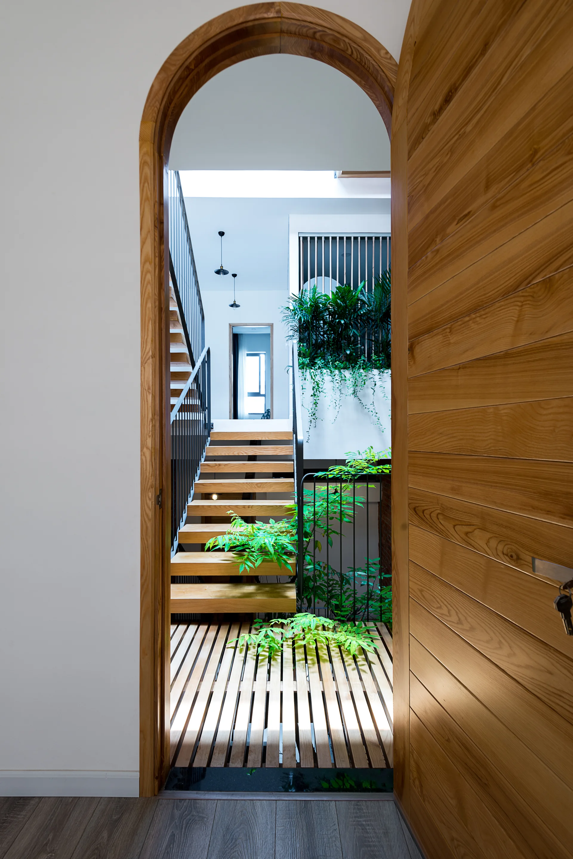 An arched wooden doorway opens to a modern staircase with floating timber treads, black metal railings, and lush indoor greenery.