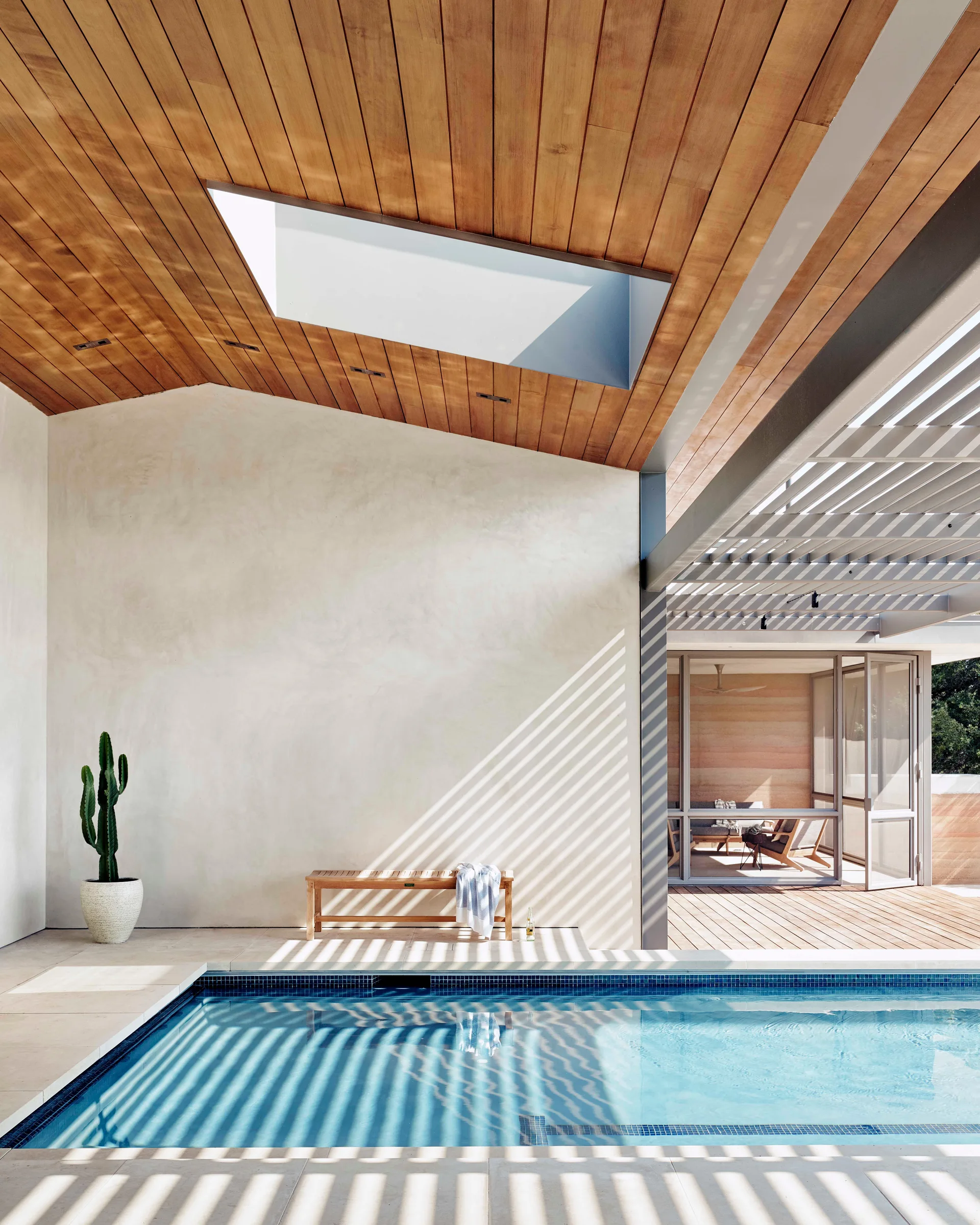 A modern outdoor swimming pool is flanked by a smooth plaster wall and a wood-plank ceiling featuring a skylight and slatted pergola shading.