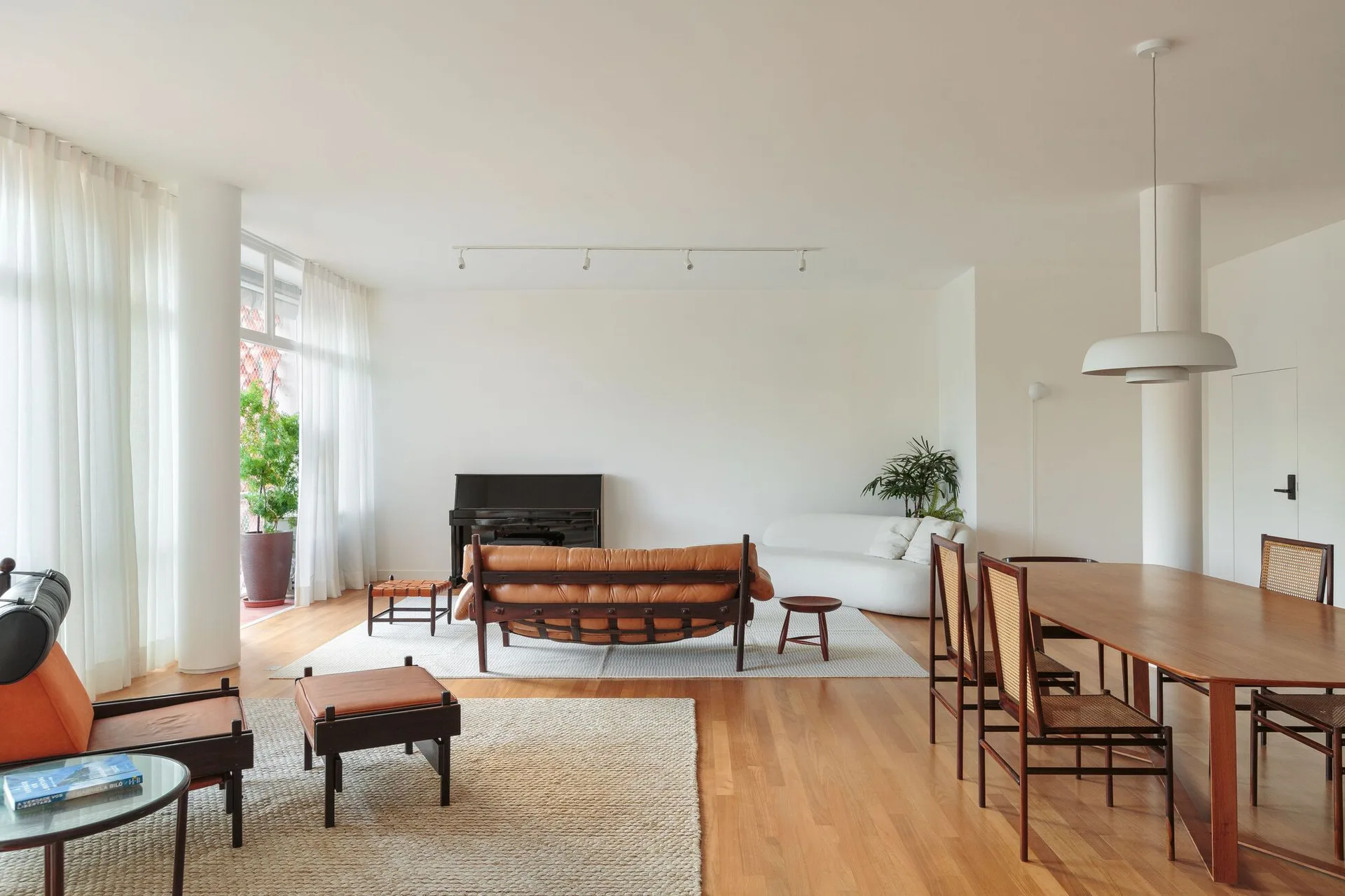 An open-plan living and dining room featuring a brown leather sofa, a wooden dining table with cane chairs, white walls, and wood floors.