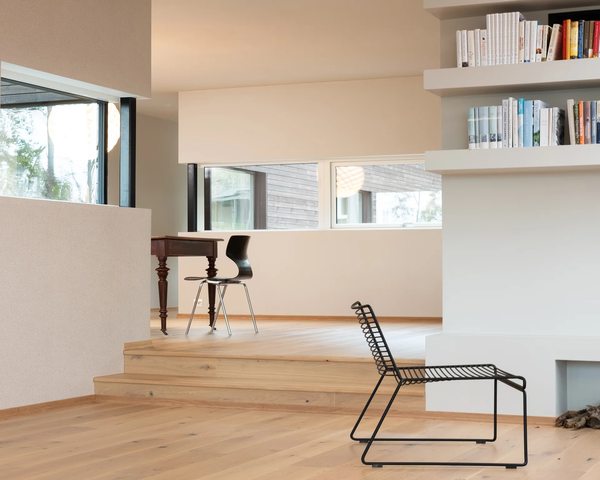A minimalist interior space featuring a multi-level wooden floor, built-in wall shelving with books, a black wire chair, and a raised platform desk.