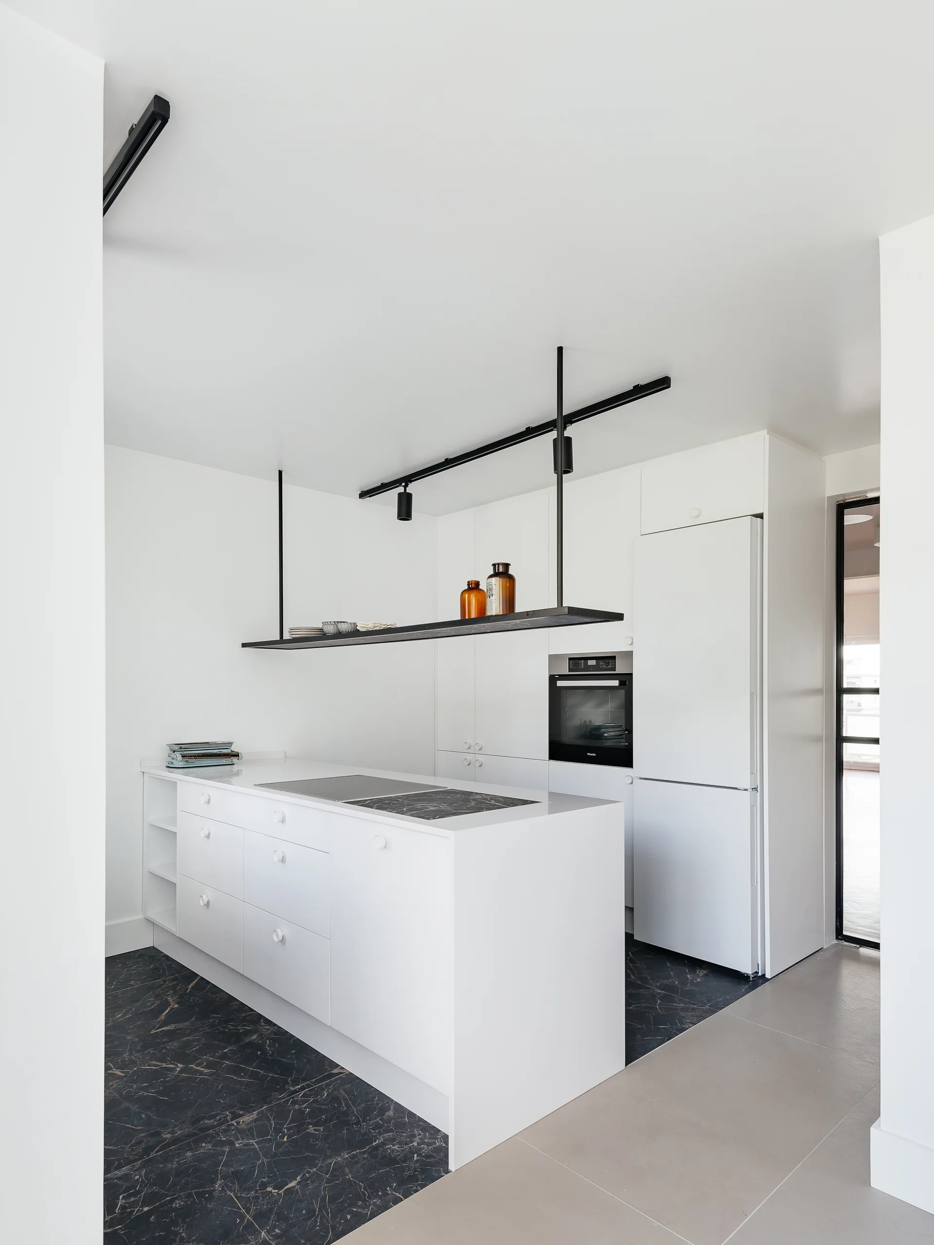 A minimalist white kitchen featuring a central island, a black suspended ceiling shelf, and dark marble floor tiling.