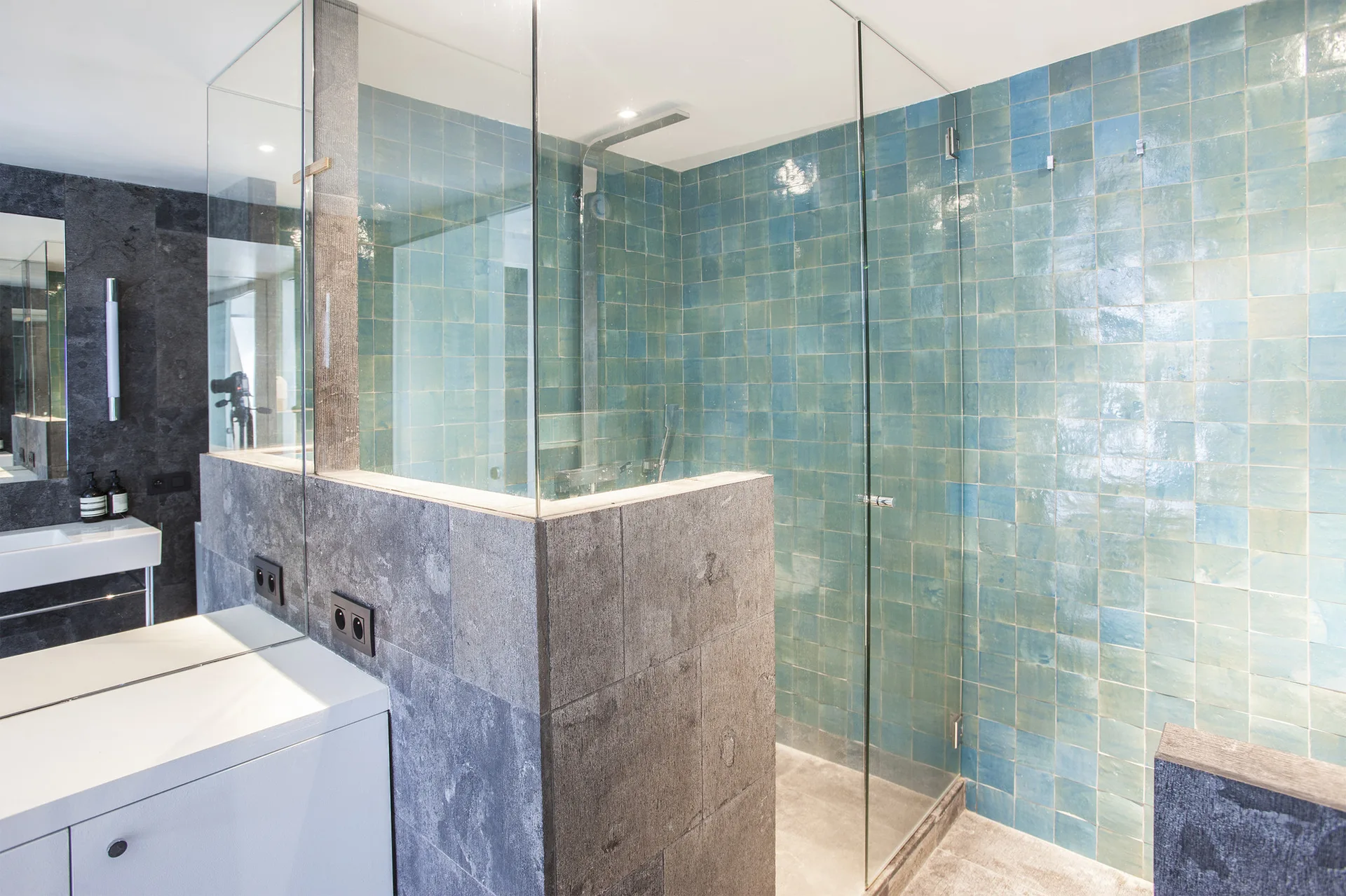 A modern bathroom features a glass shower enclosure with glossy blue square tiles, a grey textured stone half-wall, and a white vanity.
