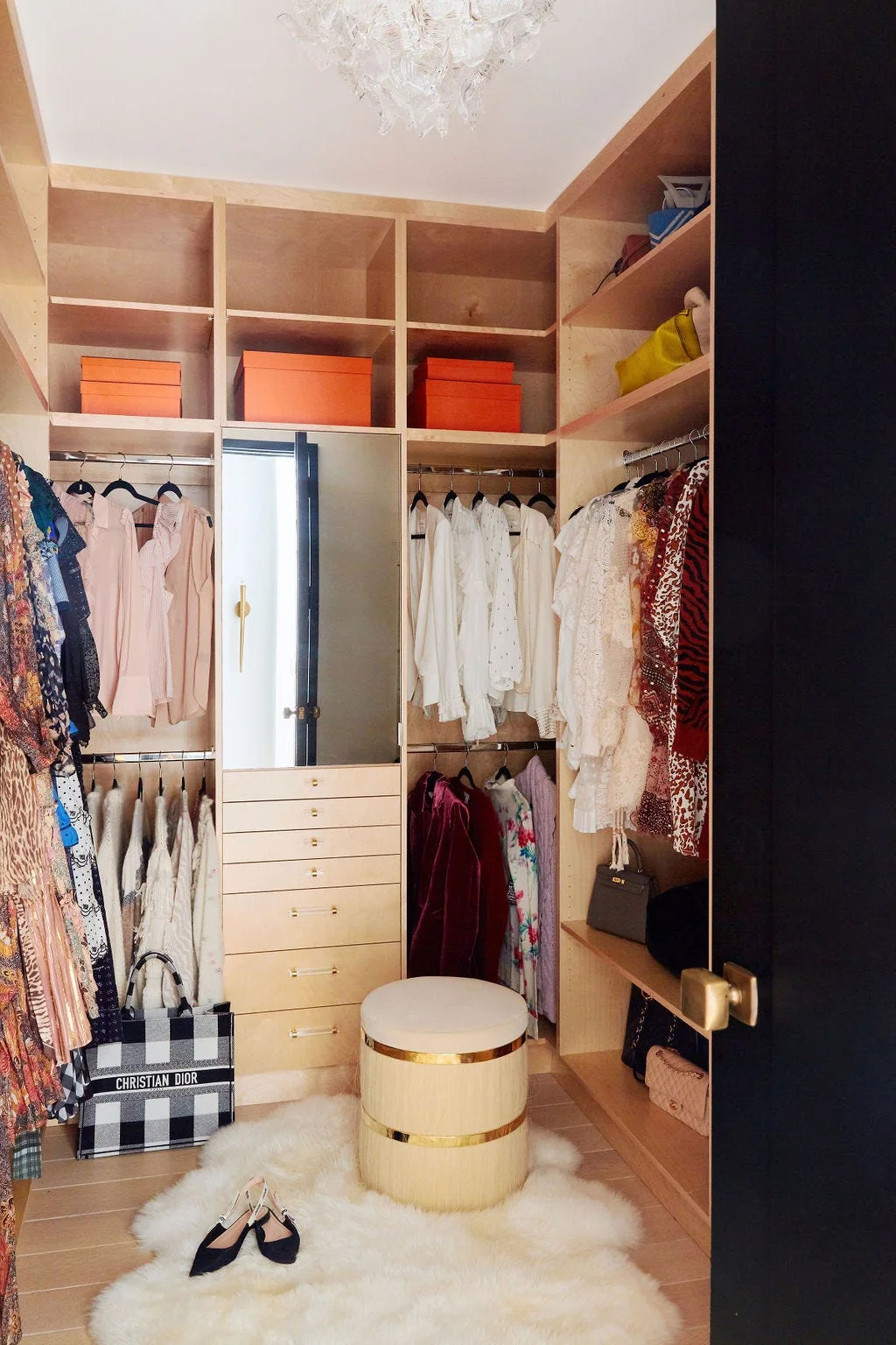 A walk-in closet with light wood shelving, clothes hanging on racks, a central drawer unit with a mirror, a round ottoman, and a white sheepskin rug.