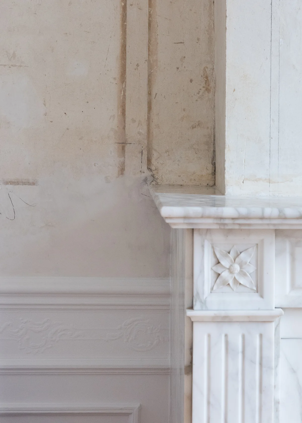 A close-up of a carved white marble fireplace mantel featuring a floral medallion next to an unfinished plaster wall with ornate wainscoting.