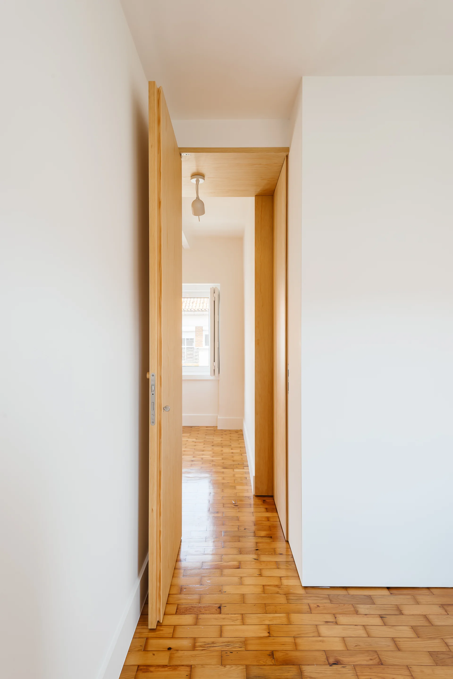 An open wooden door leads into a narrow white hallway featuring a polished parquet floor, matching wood trim, and a window at the far end.