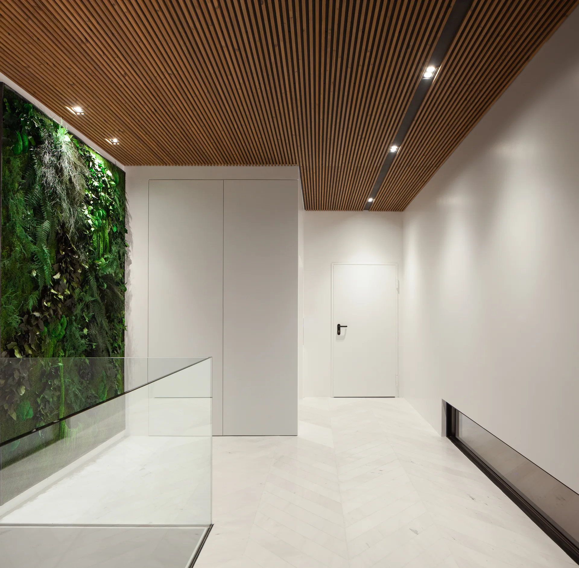 A modern hallway features a vertical garden wall, a warm wood slatted ceiling, glass railing, and white marble herringbone tile floors.