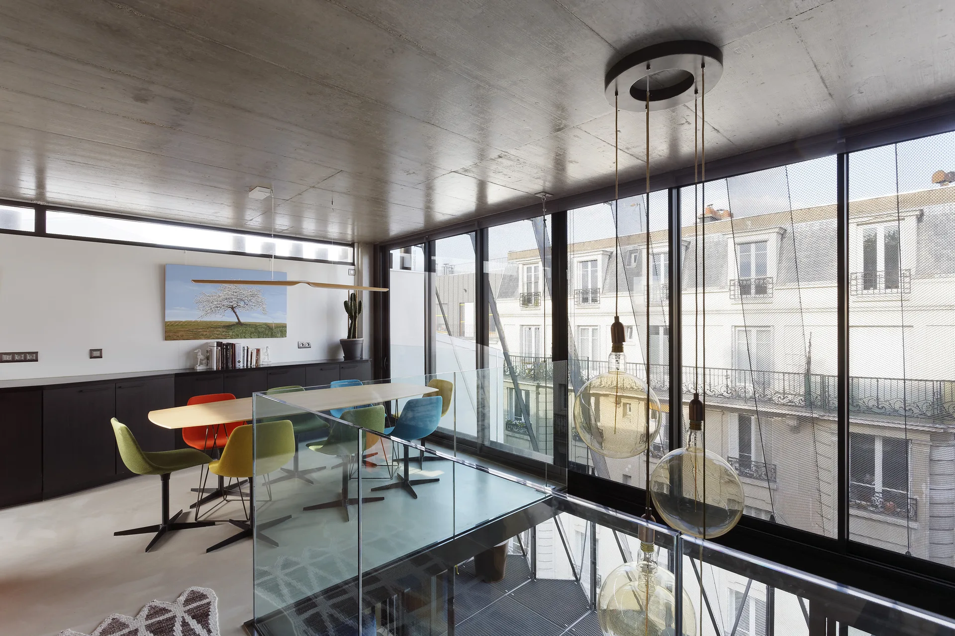 A modern loft dining area features a light wood table with colorful swivel chairs next to a glass railing, large windows, and a concrete ceiling.