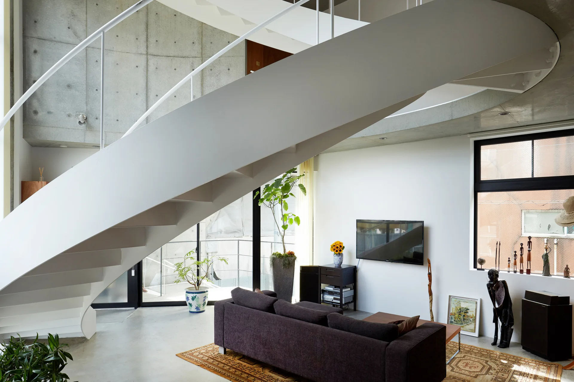 A modern living room features a curving white metal staircase, a raw concrete accent wall, and a dark sofa on a patterned rug.