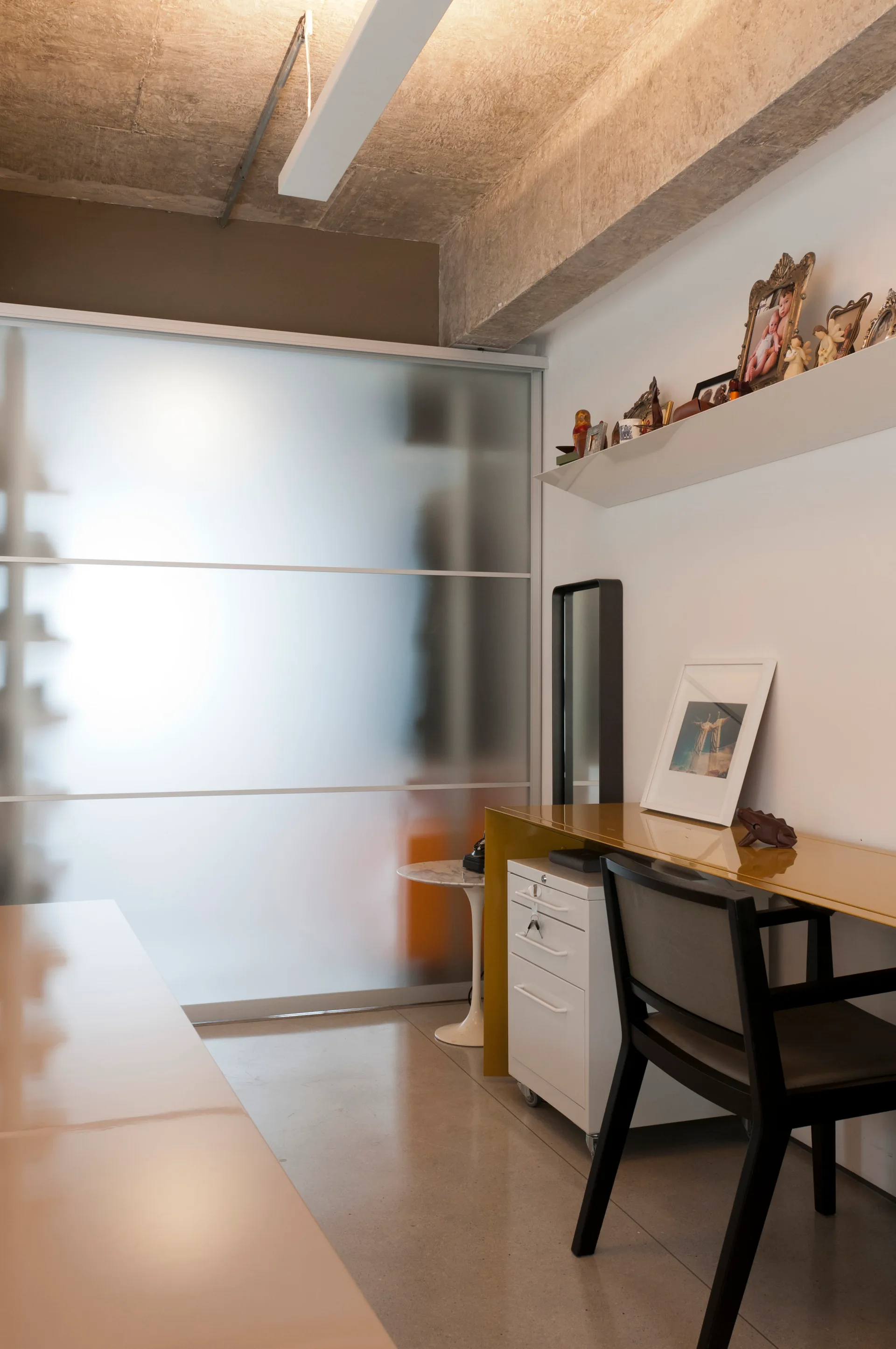 A home office workspace features a glossy yellow desk, black chair, and a frosted glass sliding door under an exposed concrete ceiling.