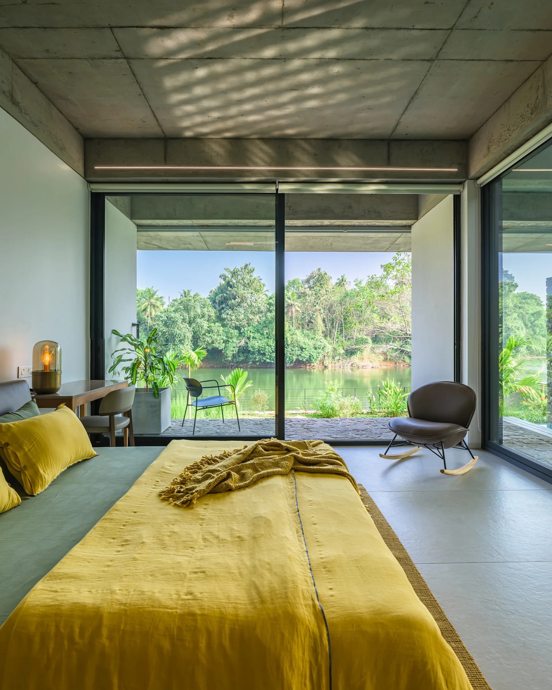 A modern bedroom features a bed with yellow bedding, a raw concrete ceiling, and sliding glass doors that look out onto a river and green trees.