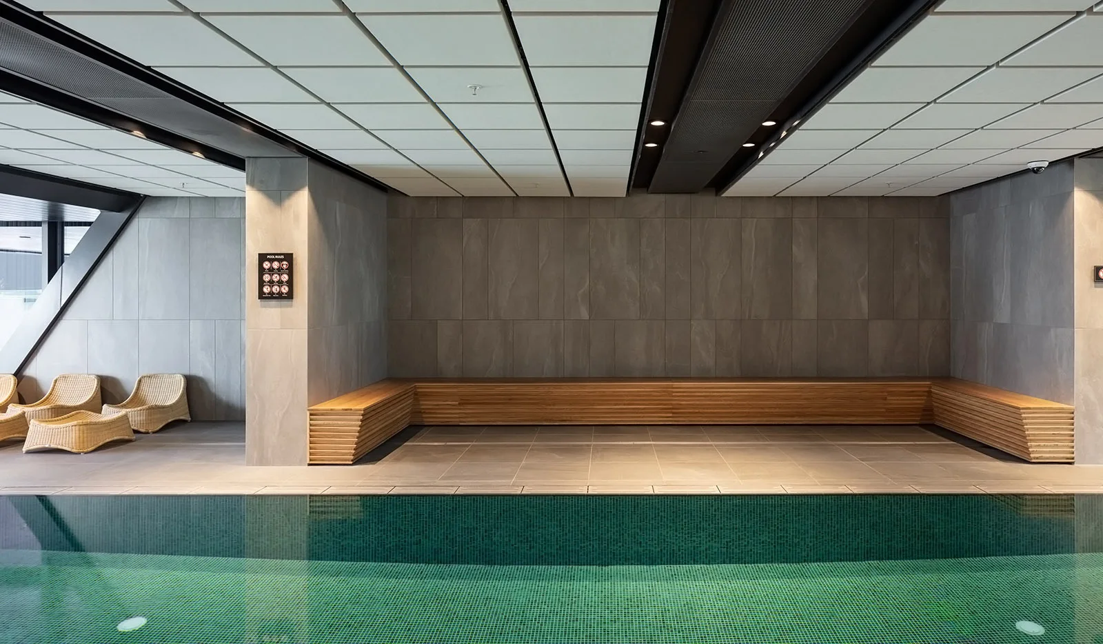 An indoor swimming pool with green mosaic tiles features grey stone wall panels and a long slatted wood bench.