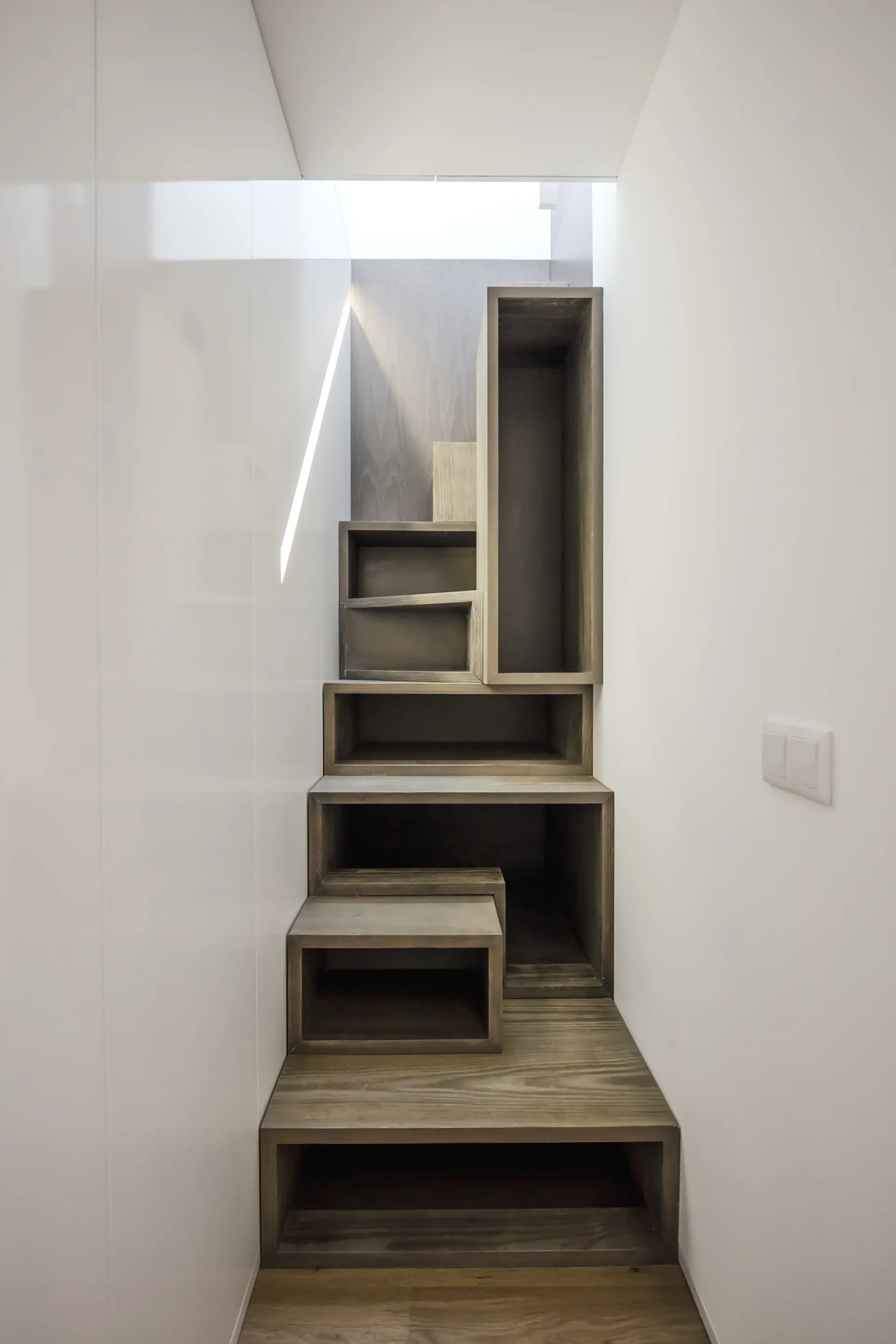 A minimalist wooden staircase with integrated open storage cubbies is built between smooth white walls under natural light from a skylight.