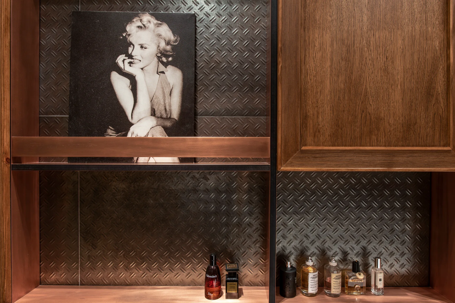 A custom wooden shelving unit with integrated lighting displays a portrait print and fragrance bottles against a dark diamond plate metal backing.