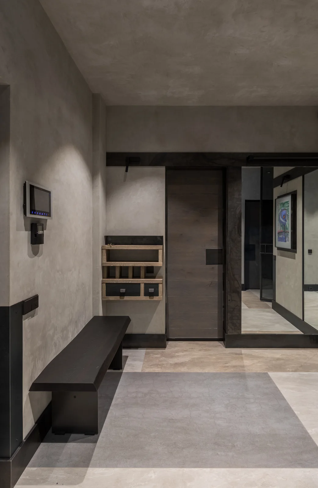 A minimalist contemporary entryway featuring textured gray walls, a dark wood bench, a small wall-mounted wooden shelf unit, and a matching door.