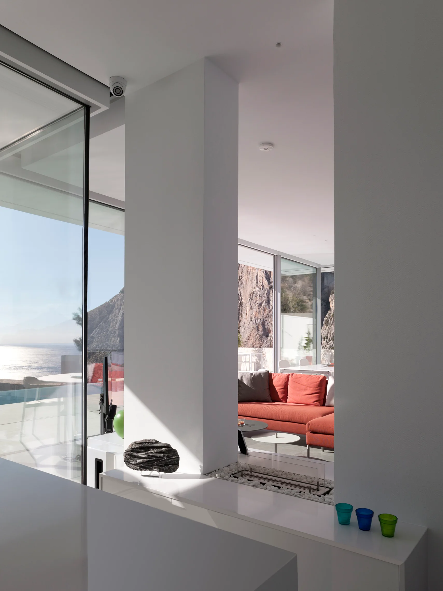 A minimalist coastal living room features a bright orange sofa and a low-profile fireplace, seen through white pillars next to floor-to-ceiling windows.
