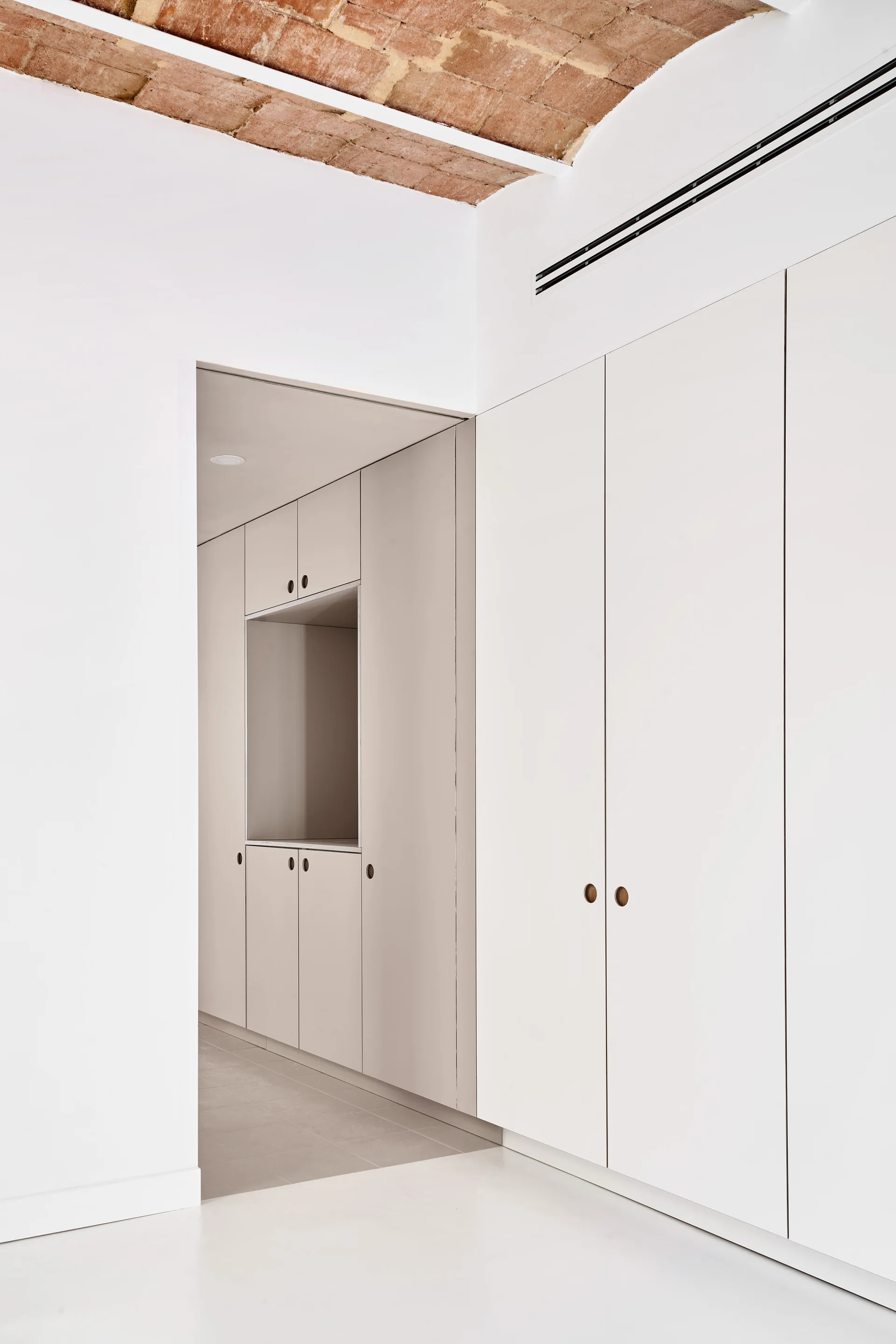 A minimalist hallway featuring custom white wardrobe cabinets with round circular cutouts beneath a rustic vaulted red brick ceiling.