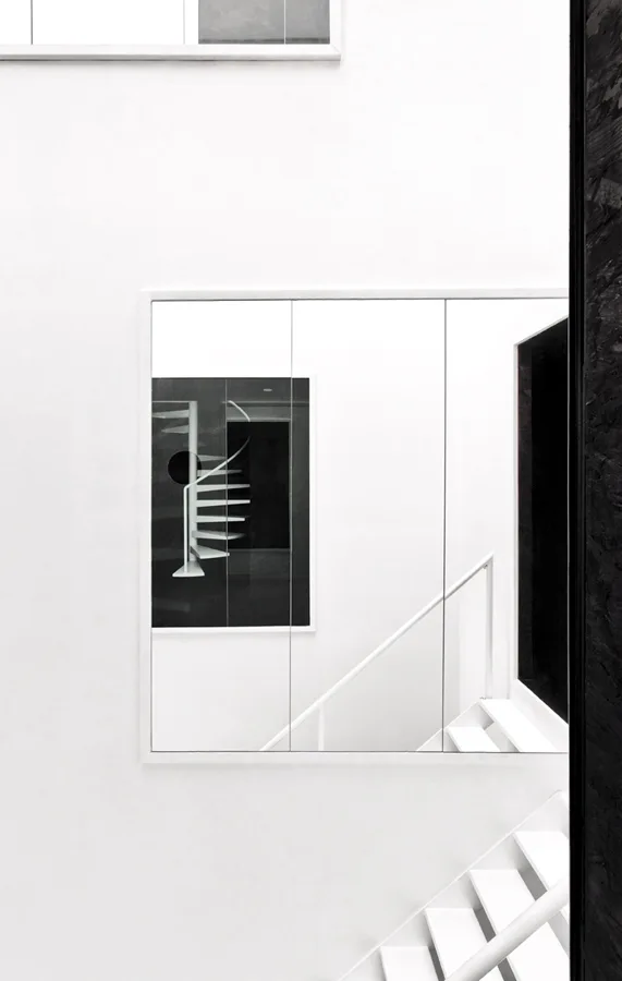 A minimalist white stairwell featuring a large three-panel mirror that reflects a white spiral staircase against a black background.
