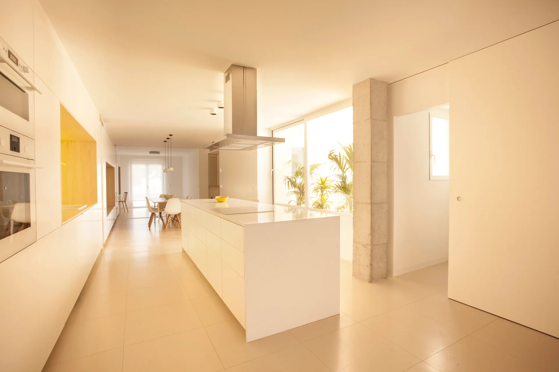 A minimalist white kitchen features a large central island with a stove hood next to an exposed concrete column and a dining area in the background.