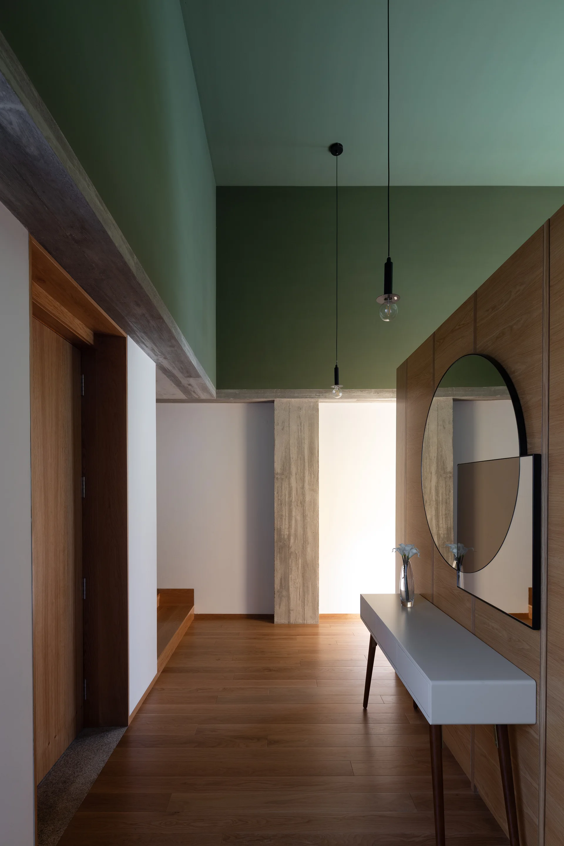 A minimalist hallway features a sage green ceiling, wood-paneled walls, a white console table with a round mirror, and an exposed concrete column.