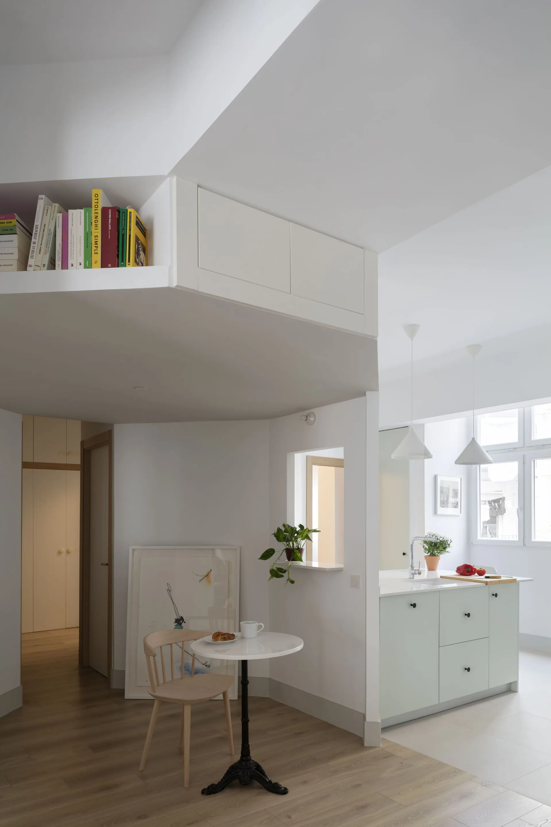 An open kitchen features a pale green island, a small round white bistro table with a wooden chair, and an overhead bookshelf near the ceiling.