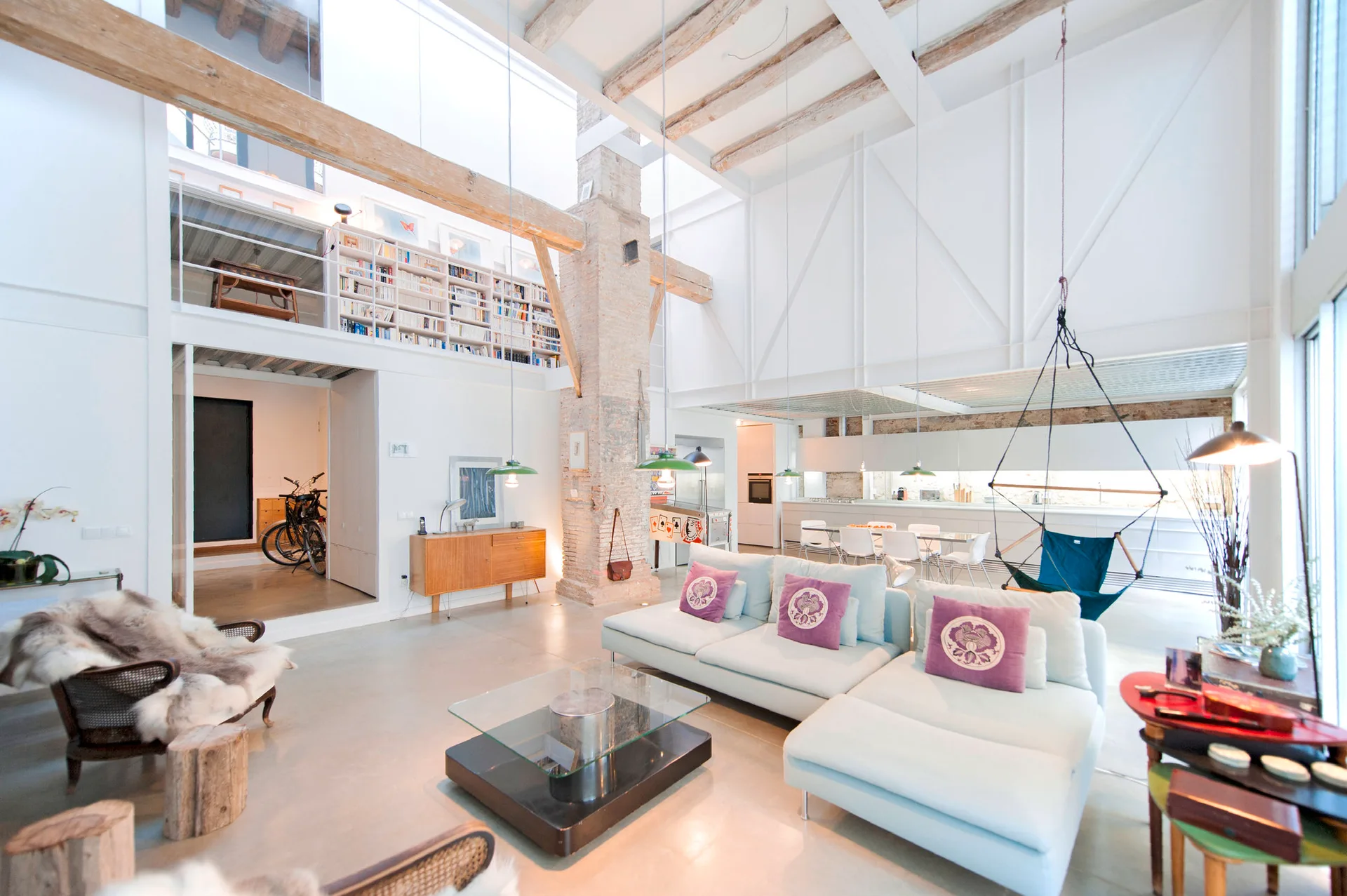 An open-plan loft living room features a light blue modular sofa, a tall brick column, exposed wooden ceiling beams, and a mezzanine bookcase.