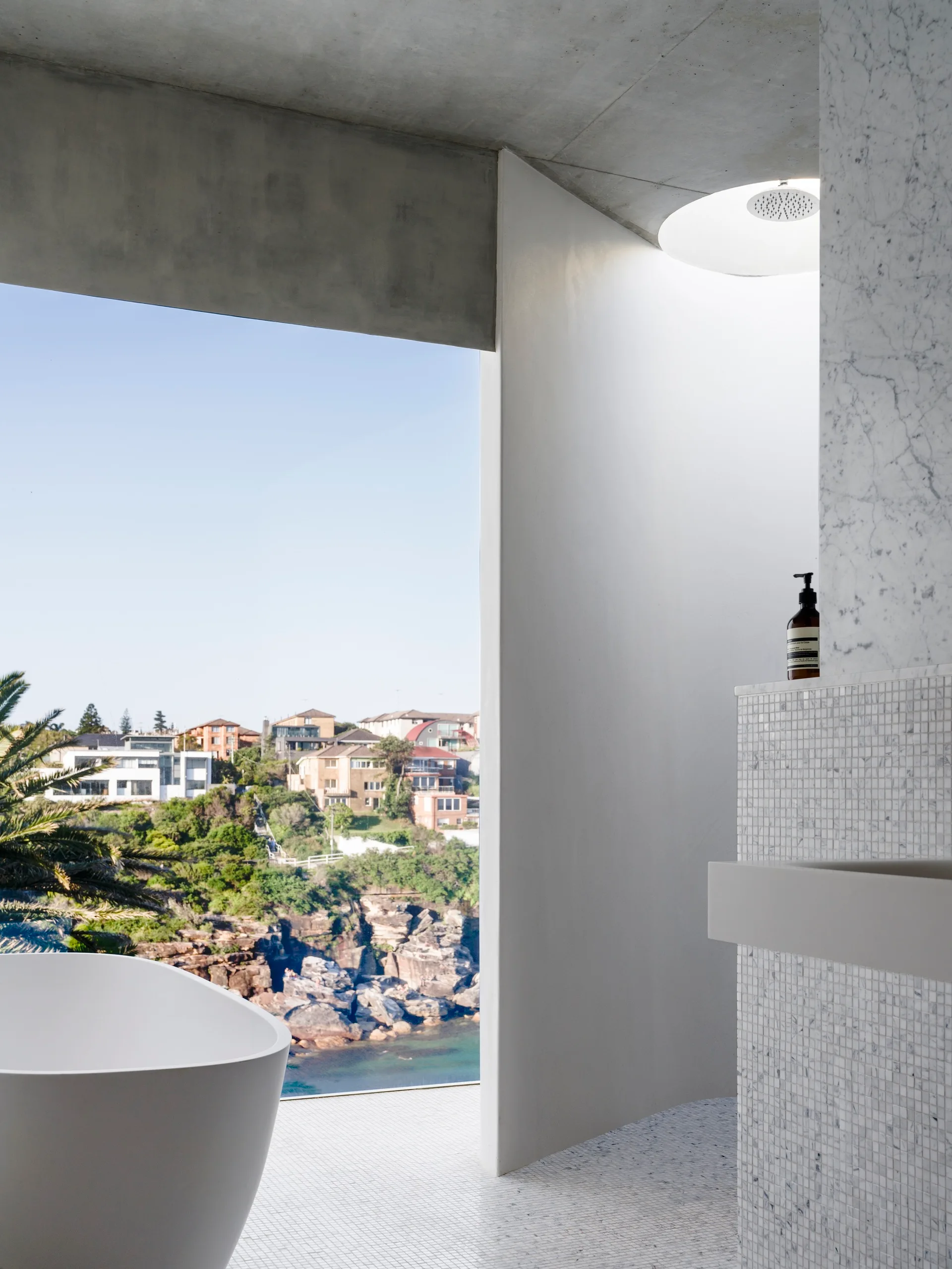 A minimalist bathroom featuring a white freestanding bathtub next to a large window overlooking a rocky coastal landscape under a grey concrete ceiling.