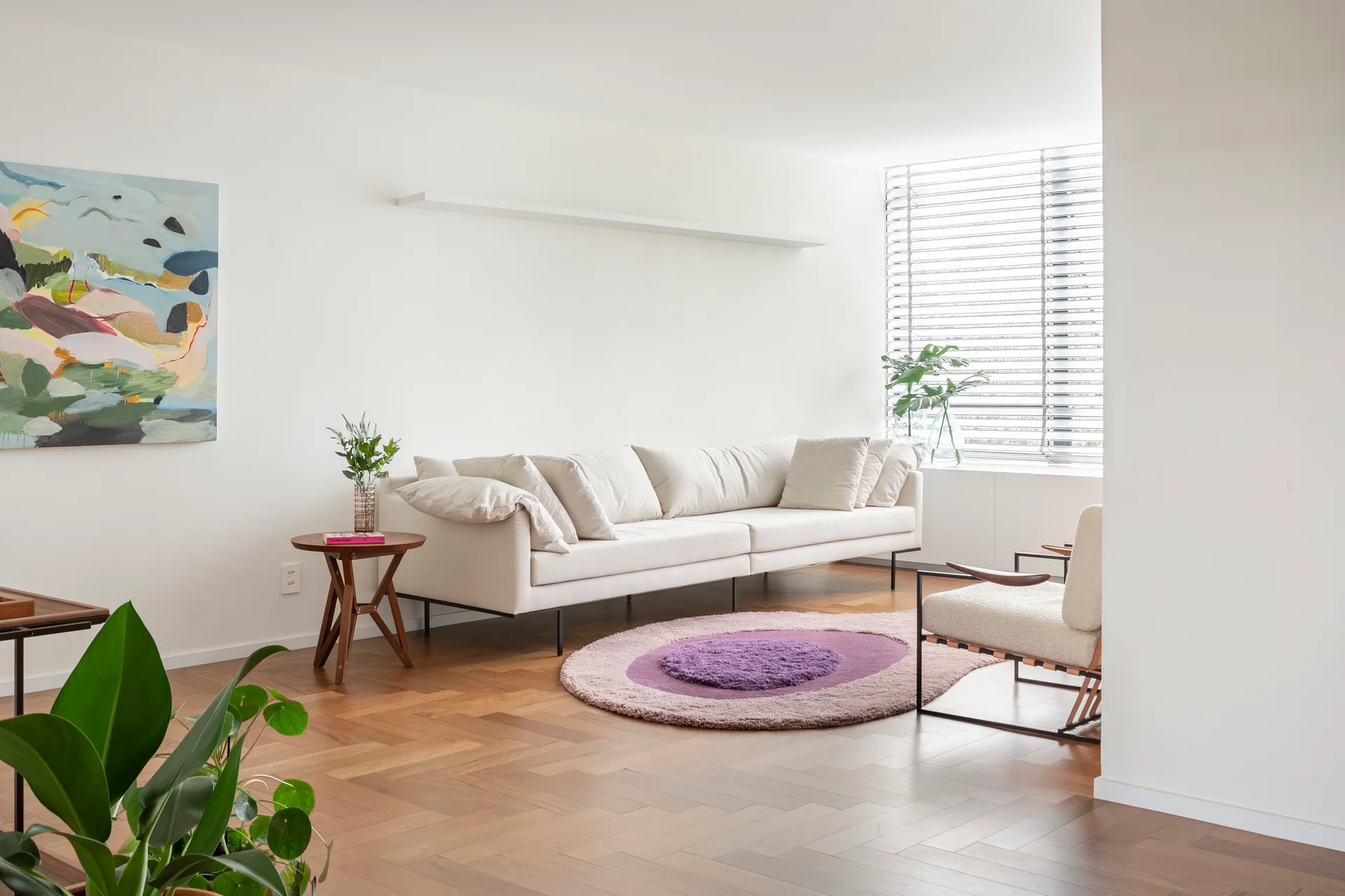 A bright living room features a cream sofa, a circular purple rug on parquet wood flooring, and a large window with blinds.