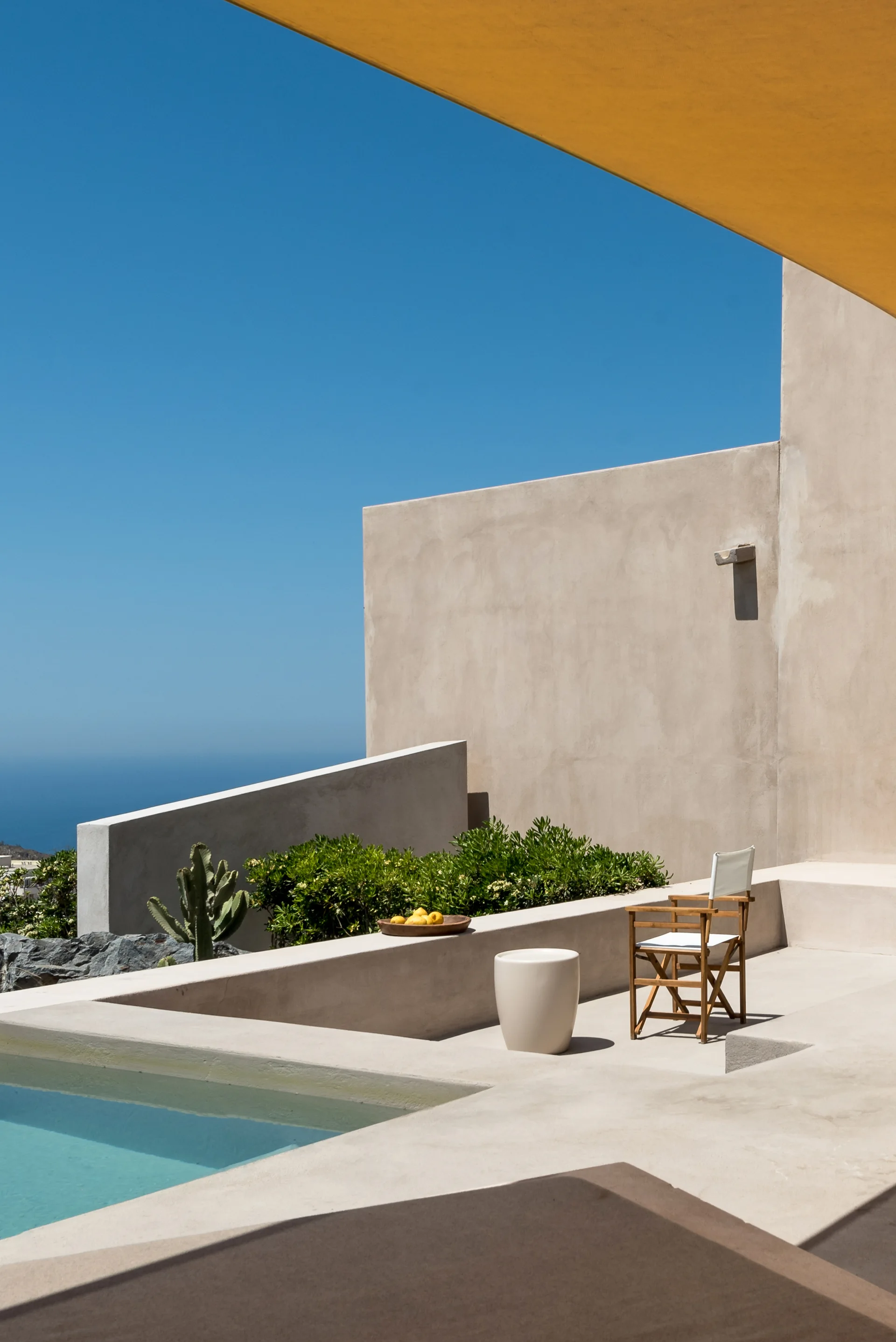 A minimalist concrete patio features a small swimming pool, a wooden folding director's chair, a low round side table, and a yellow awning overhead.
