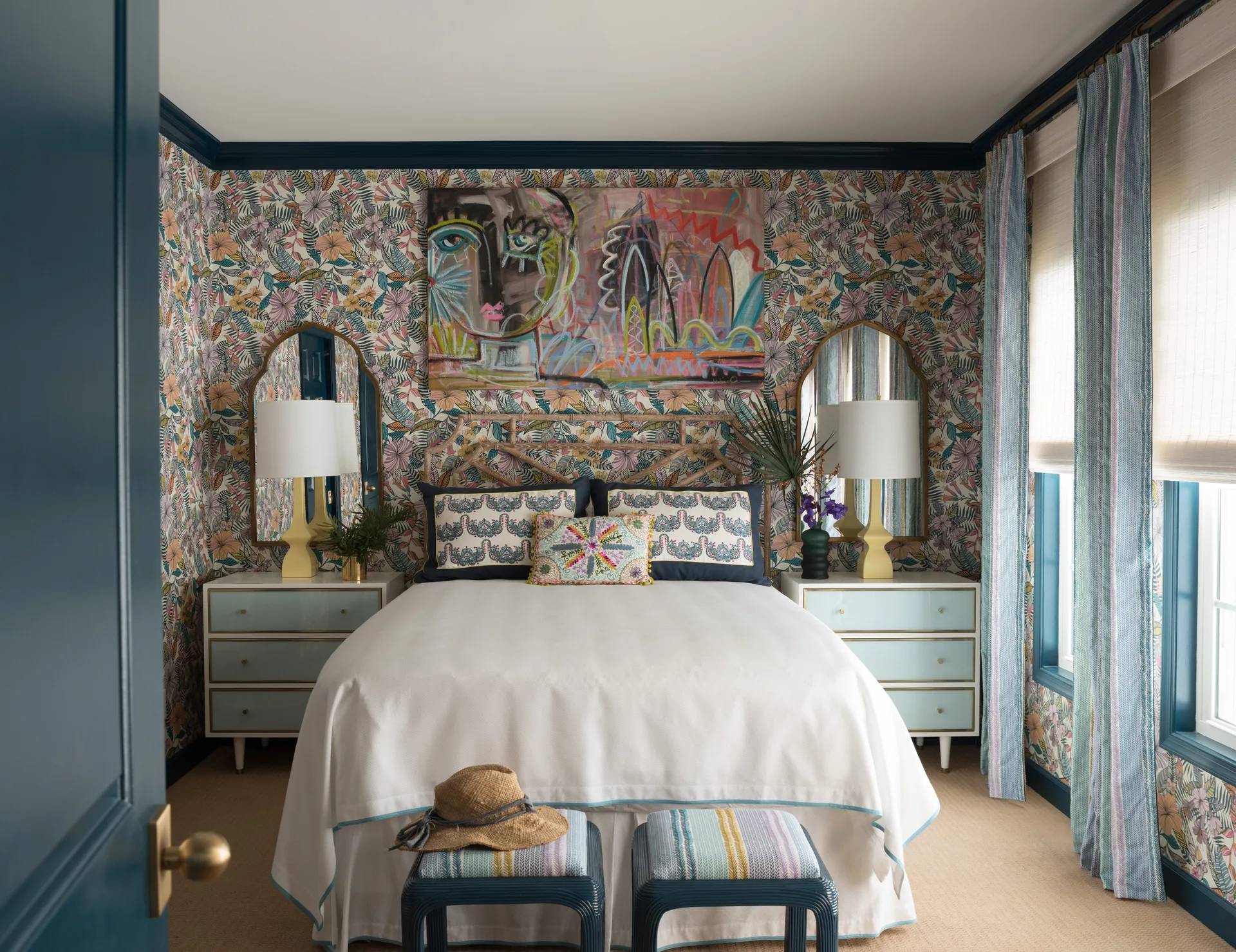 A maximalist bedroom featuring colorful floral wallpaper, a bed with a rattan headboard, and matching light blue nightstands with gold arched mirrors.