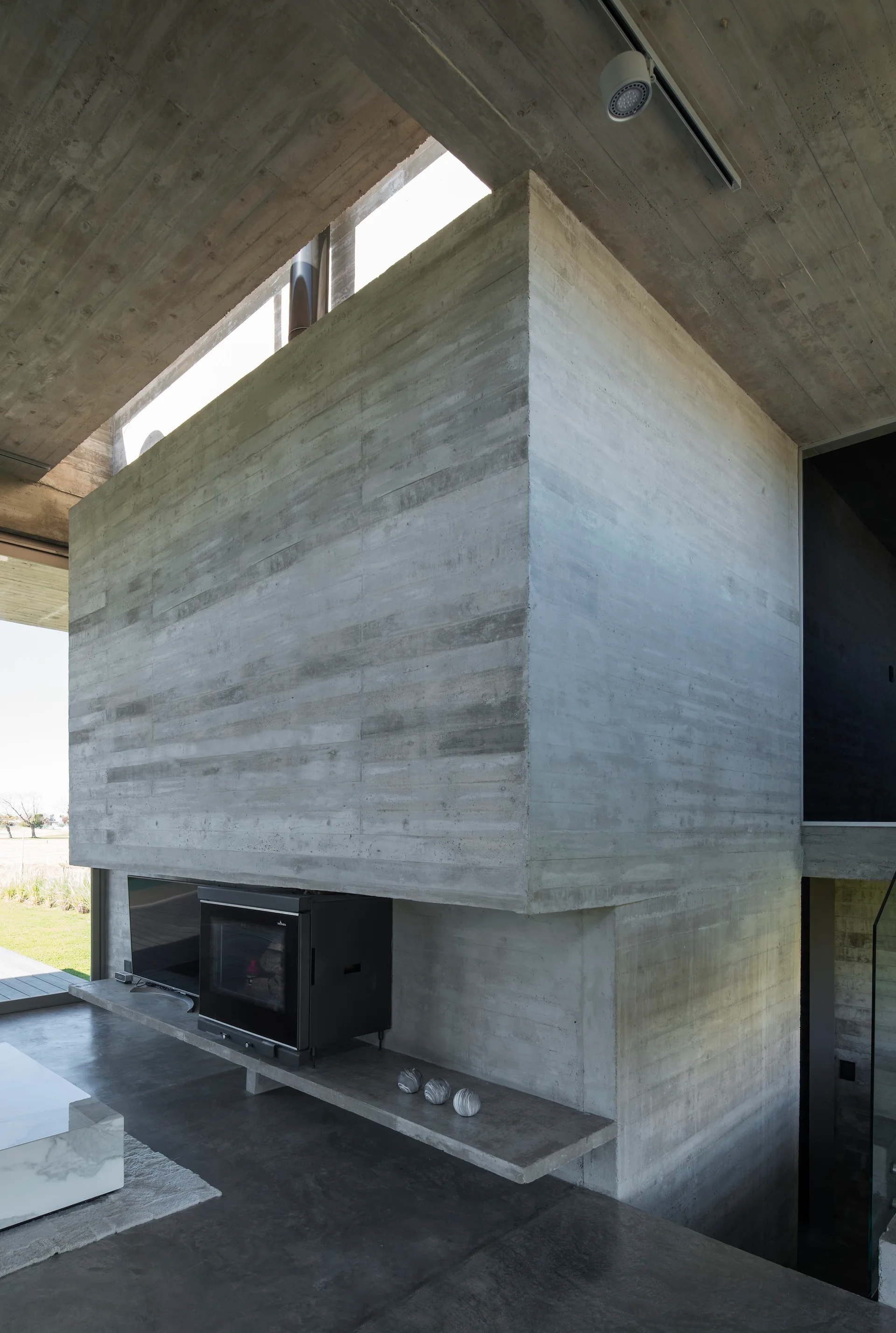 A massive board-marked concrete wall houses a black metal fireplace and TV under a concrete ceiling with a narrow clerestory window.