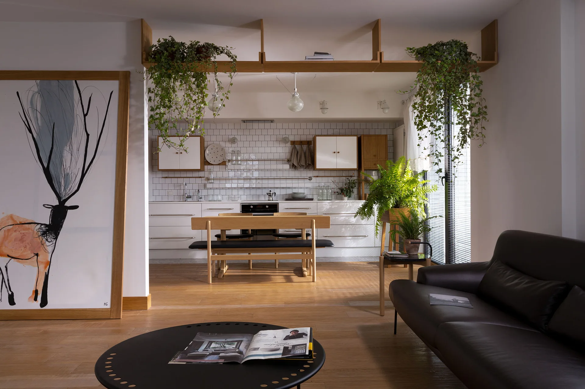 An open plan kitchen and living space features white subway tile, a light wood dining set, a black leather sofa, and hanging green plants on a high shelf.
