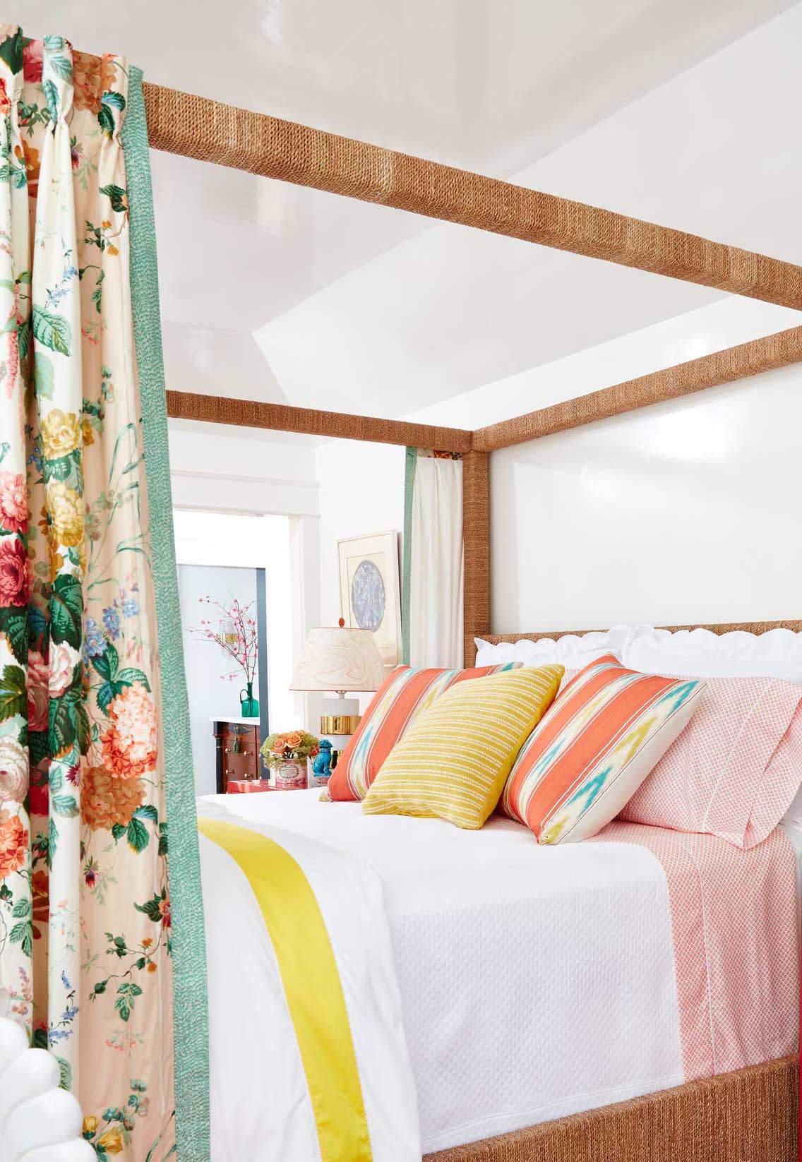 A bedroom features a woven four-poster bed frame dressed with orange and yellow striped pillows and colorful floral curtains.