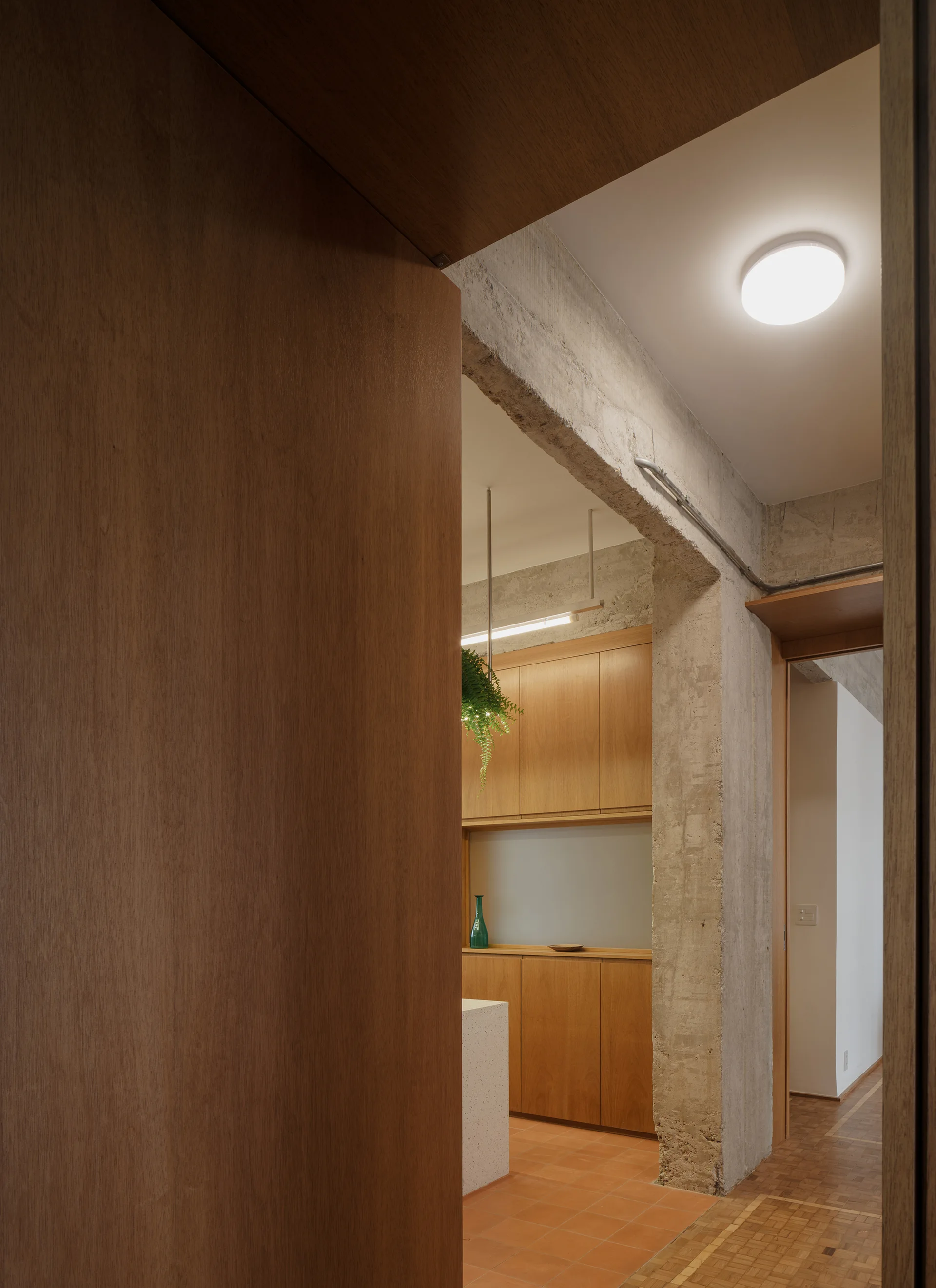 A view through a wooden doorway showing a kitchen with warm wood cabinets, terracotta tile flooring, and a rough exposed concrete column.