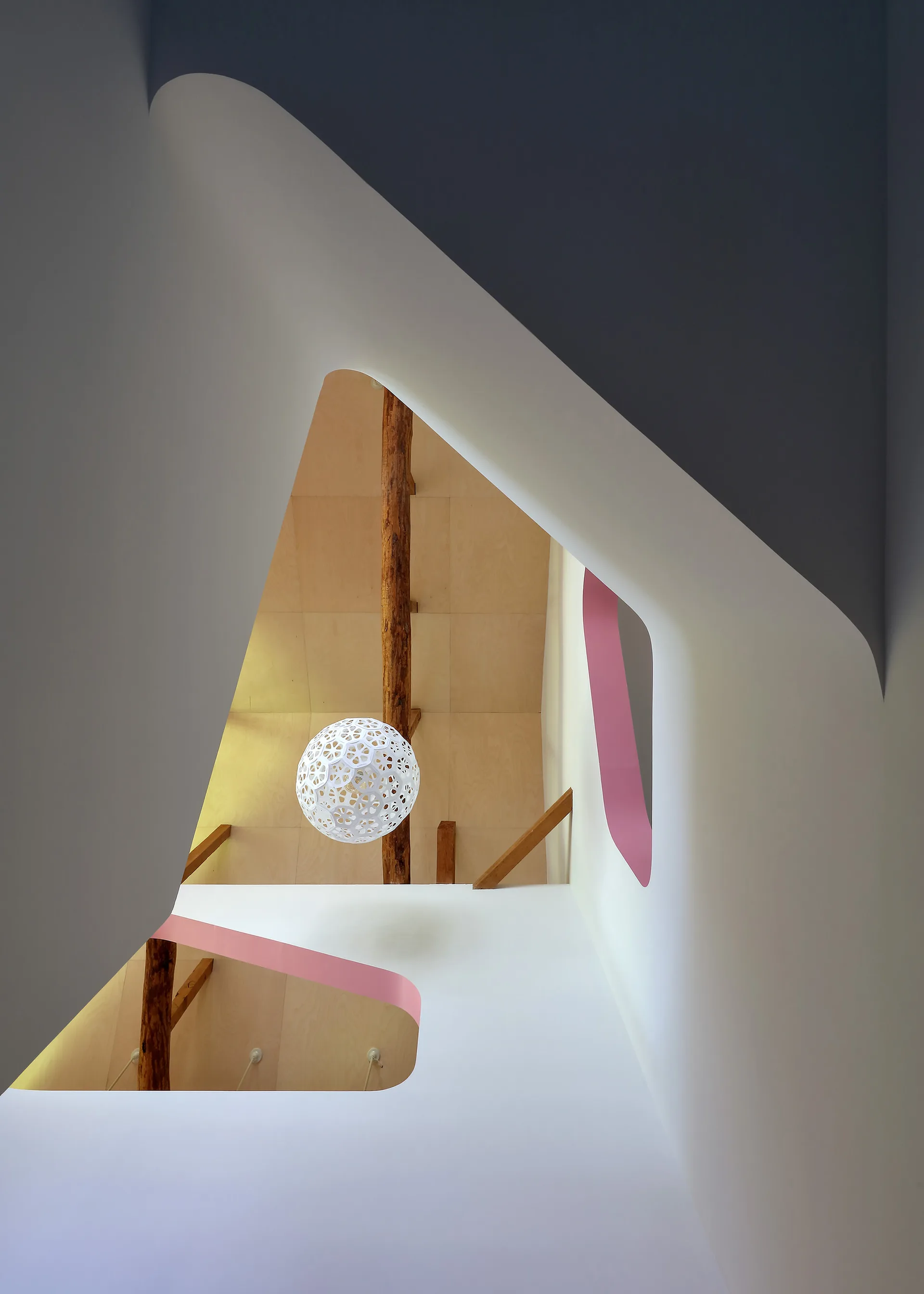 Looking up through organic triangular wall openings with pink trim to reveal a plywood ceiling, rustic wooden beams, and a white patterned pendant light.
