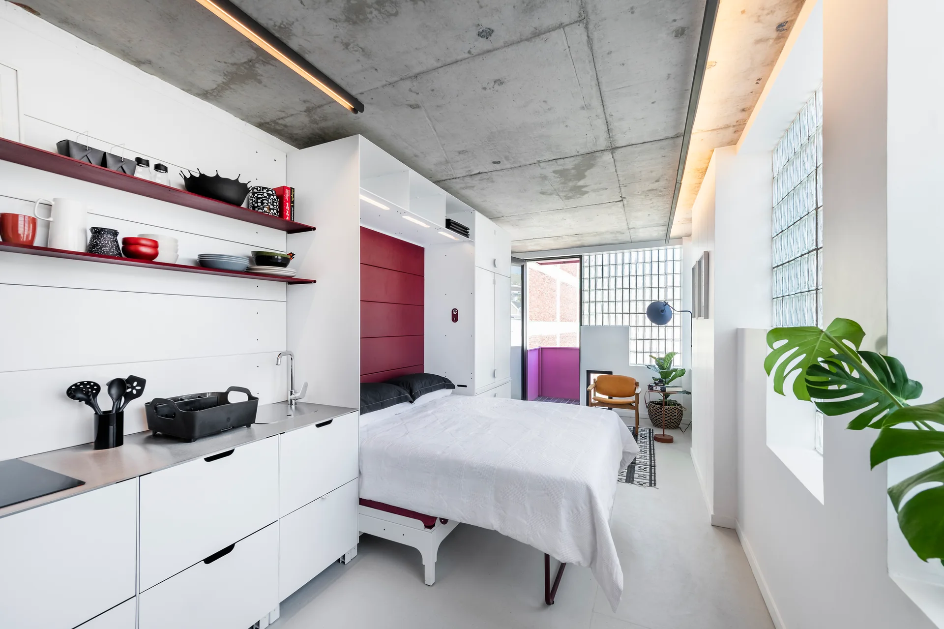 A modern studio apartment features a built-in bed and white kitchenette under an exposed concrete ceiling with glass block windows.