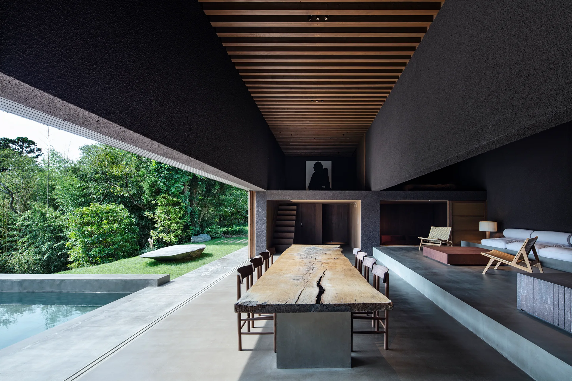 An indoor-outdoor dining space features a live-edge wood table, concrete floors, a slatted ceiling, and an open wall leading to a pool.