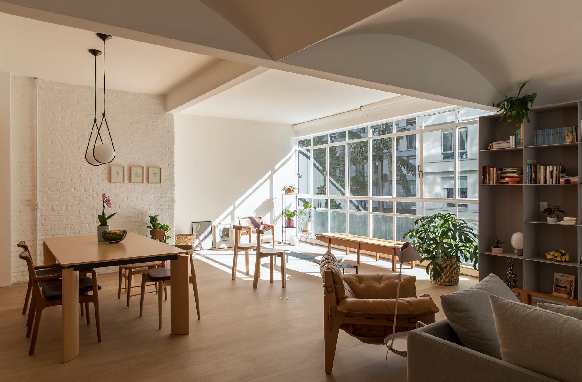 A bright open-plan living and dining space features a wooden dining table, white painted brick wall, and a large multi-pane window.